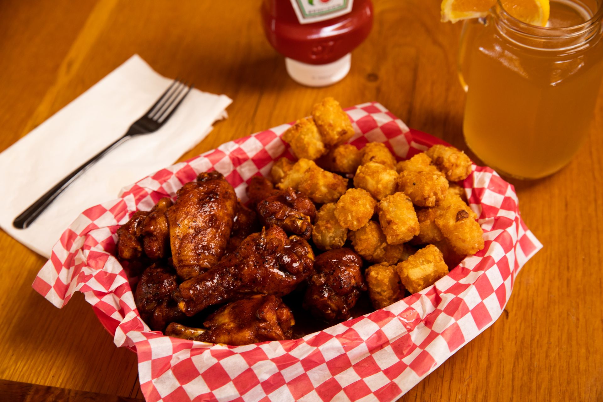 A basket of chicken wings and tots next to a bottle of ketchup