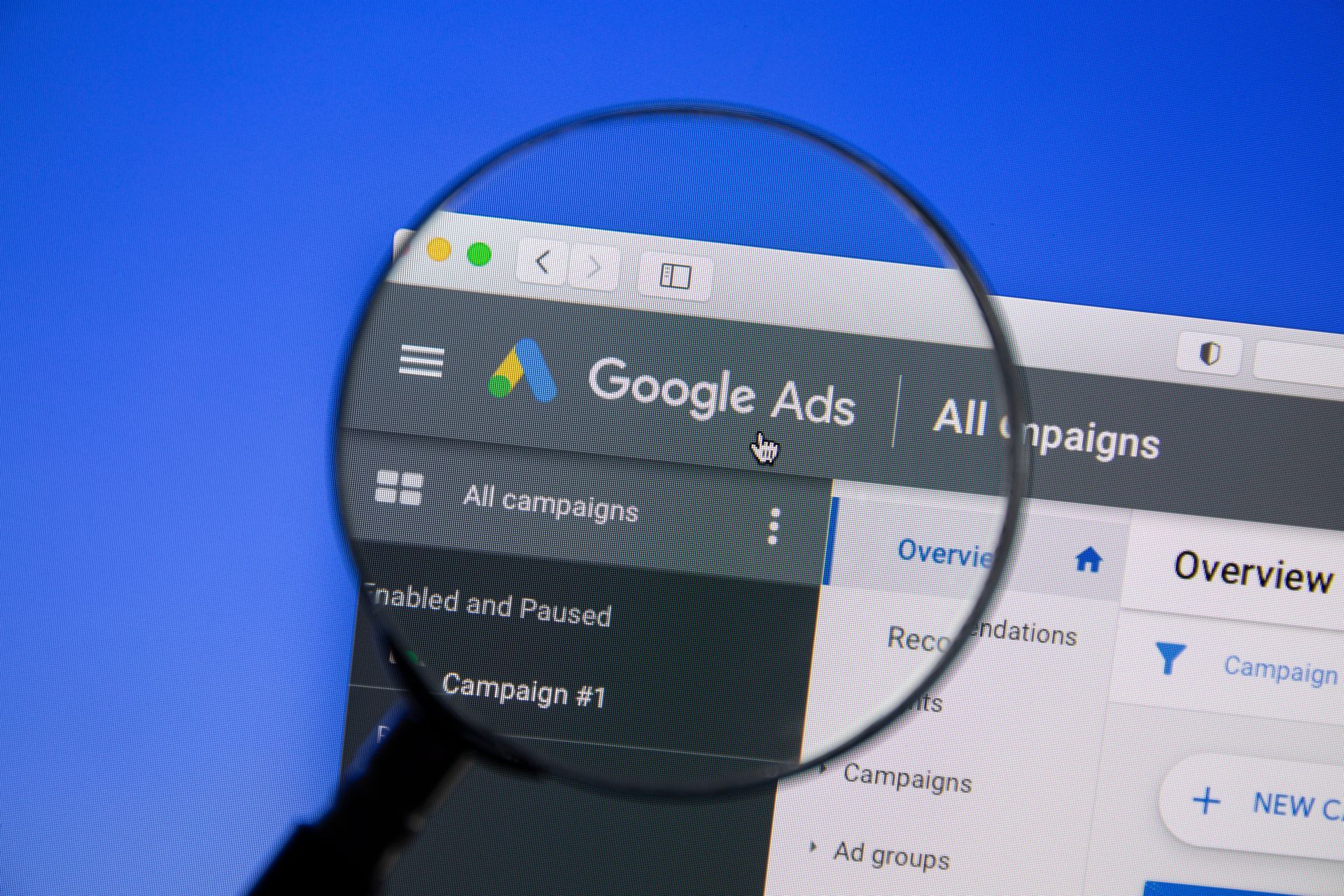 Magnifying glass over a laptop showing Google Ads dashboard in dark mode, with overview panel visible