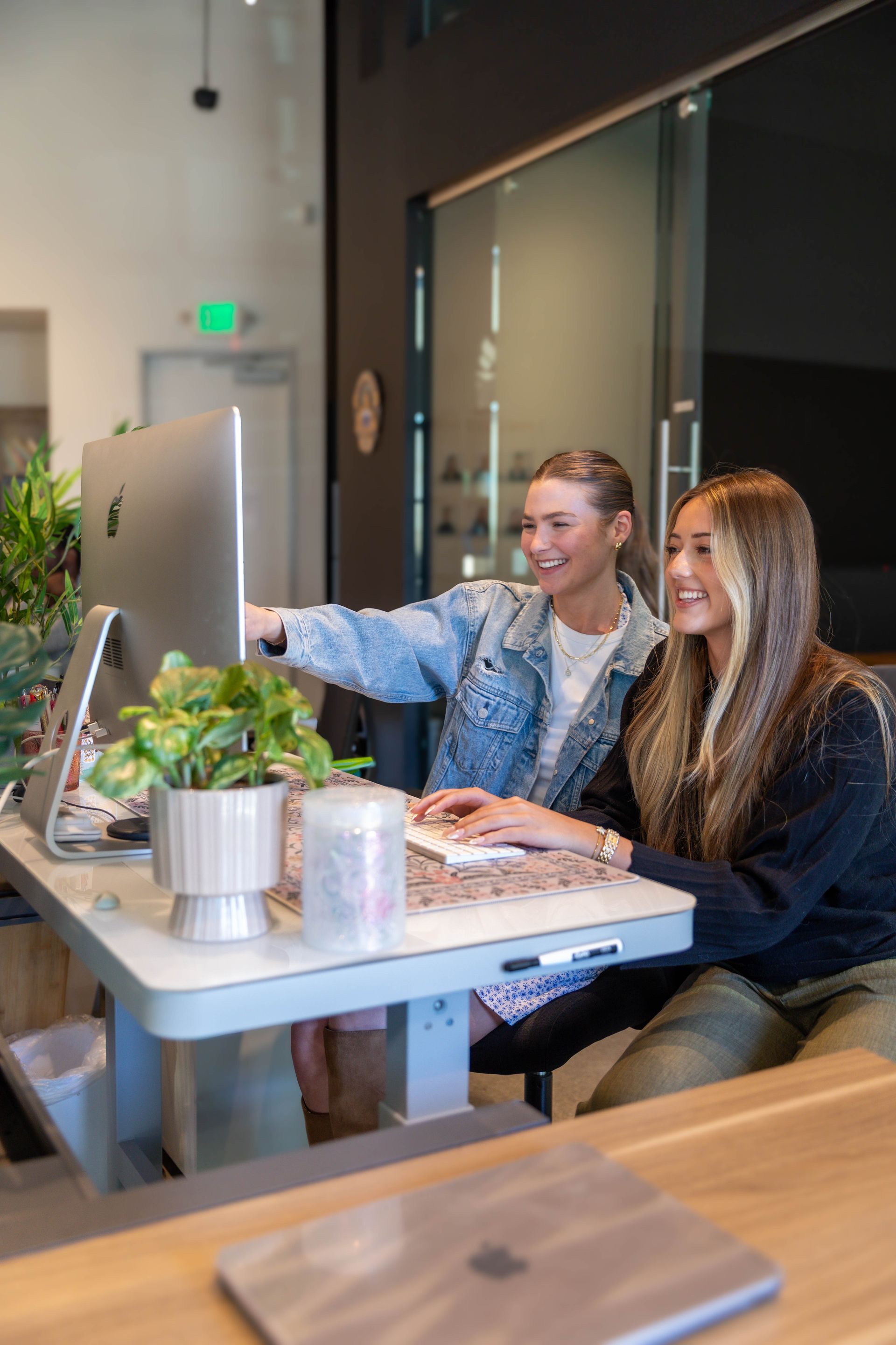 Two smiling young adults in an office setting; man with beard, woman with long hair, both looking at the camera.