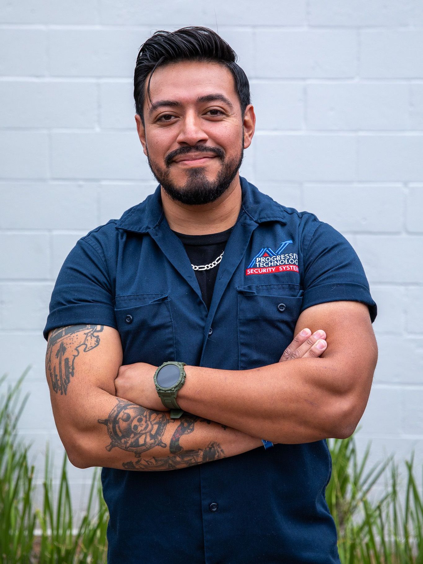 A man with a beard and tattoos is standing with his arms crossed in front of a white brick wall.
