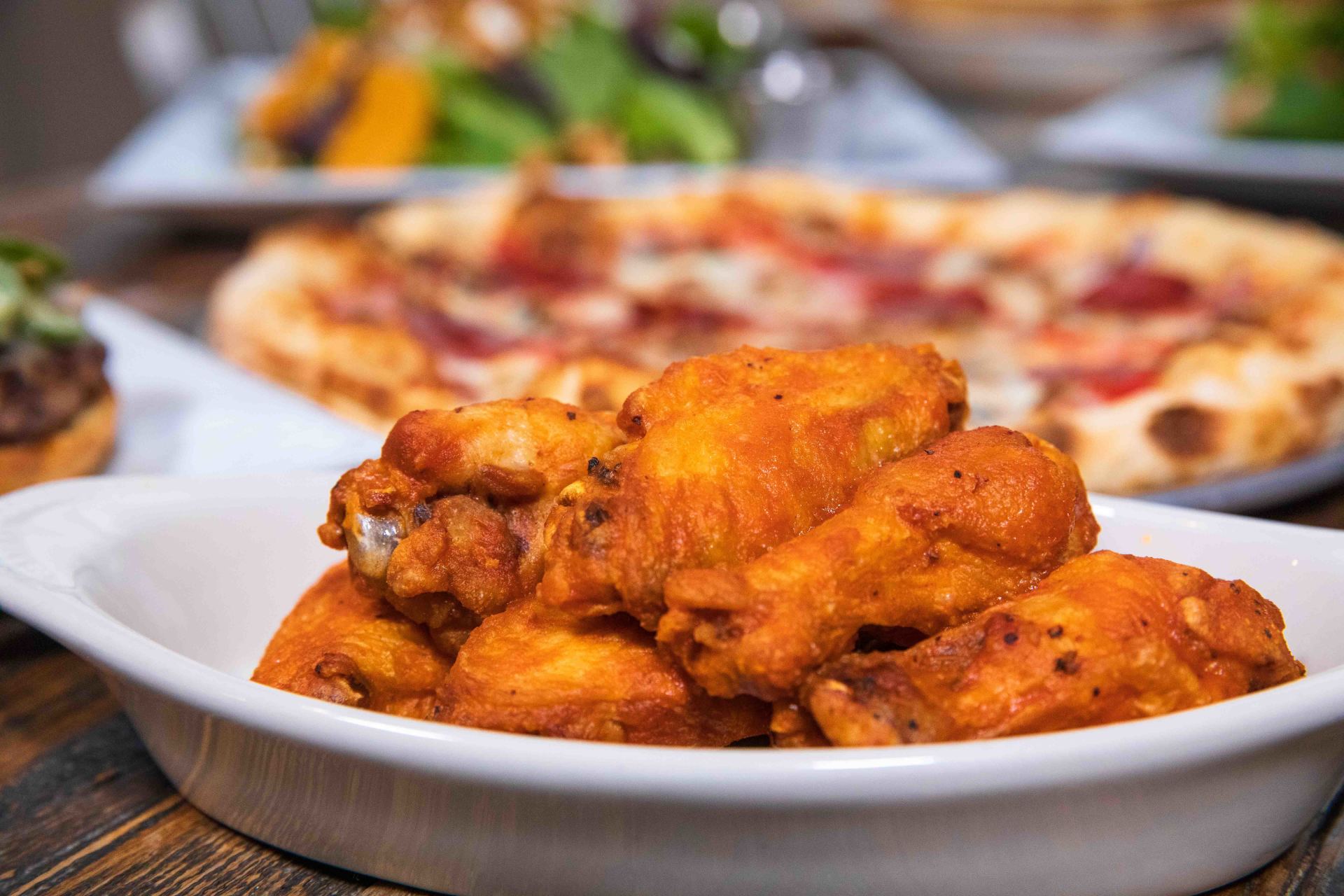 A bowl of chicken wings with a pizza in the background