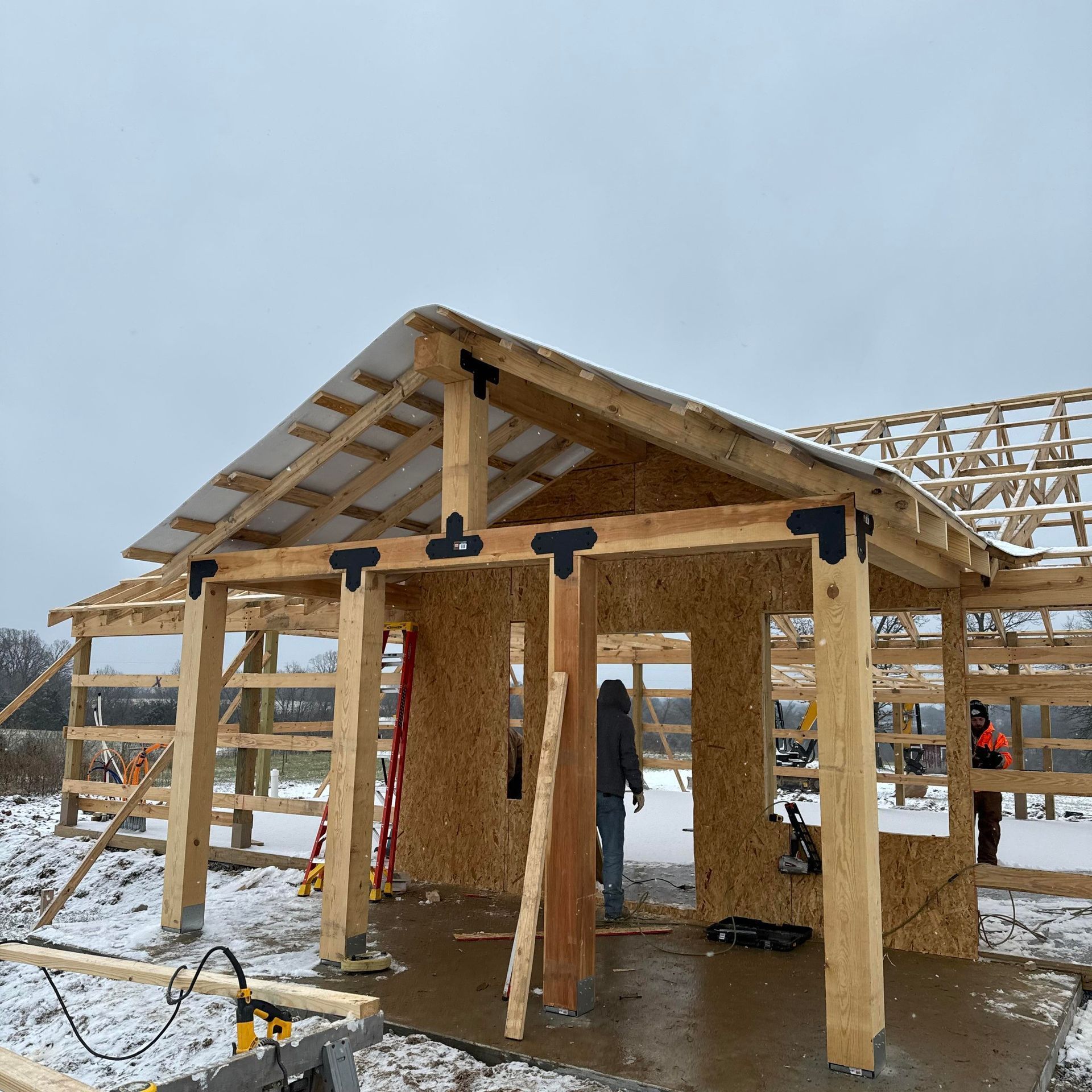 A construction site shows the timber frame and OSB sheathing of a house exterior during a snowy winter day.