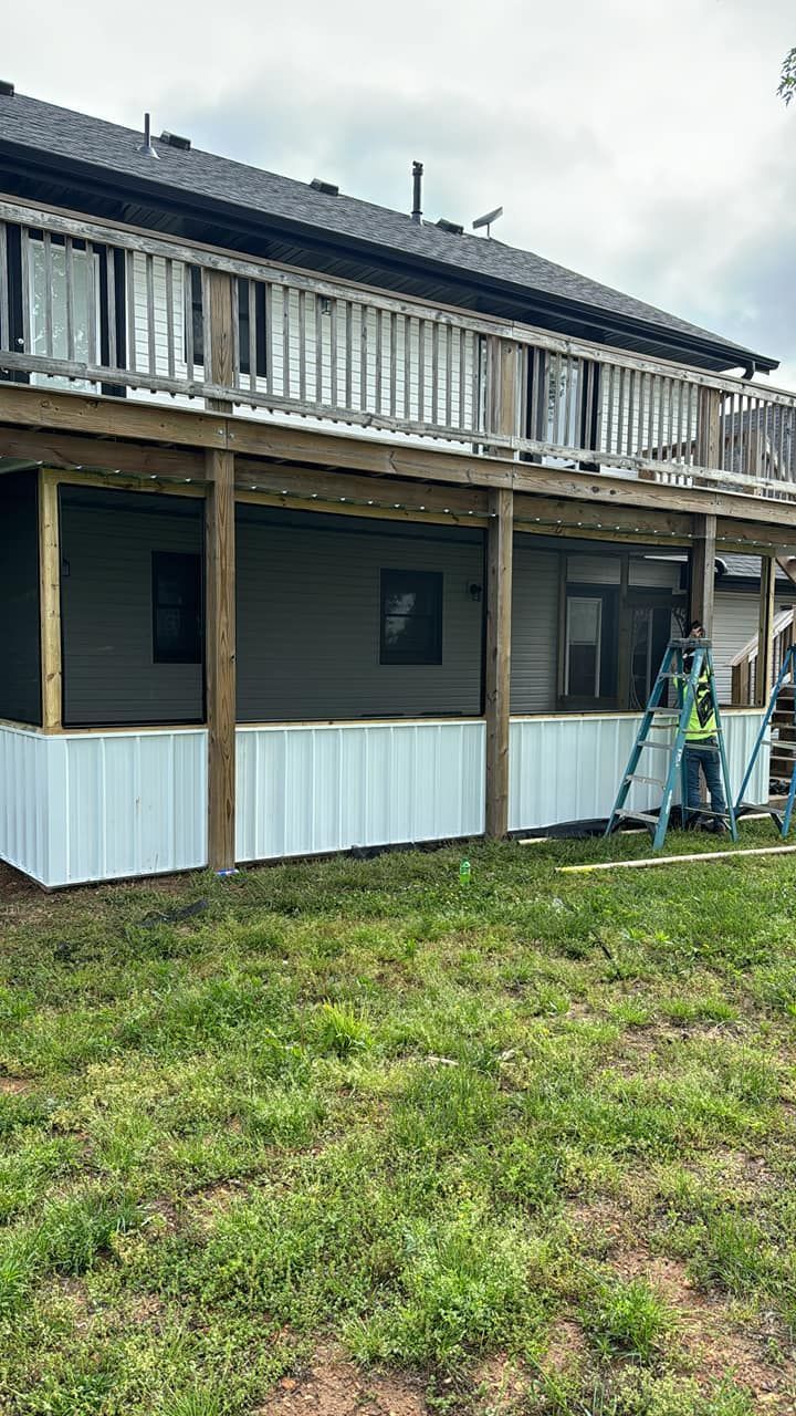 A construction worker on a ladder installing white vertical siding under a wood-framed deck attached to a gray house.