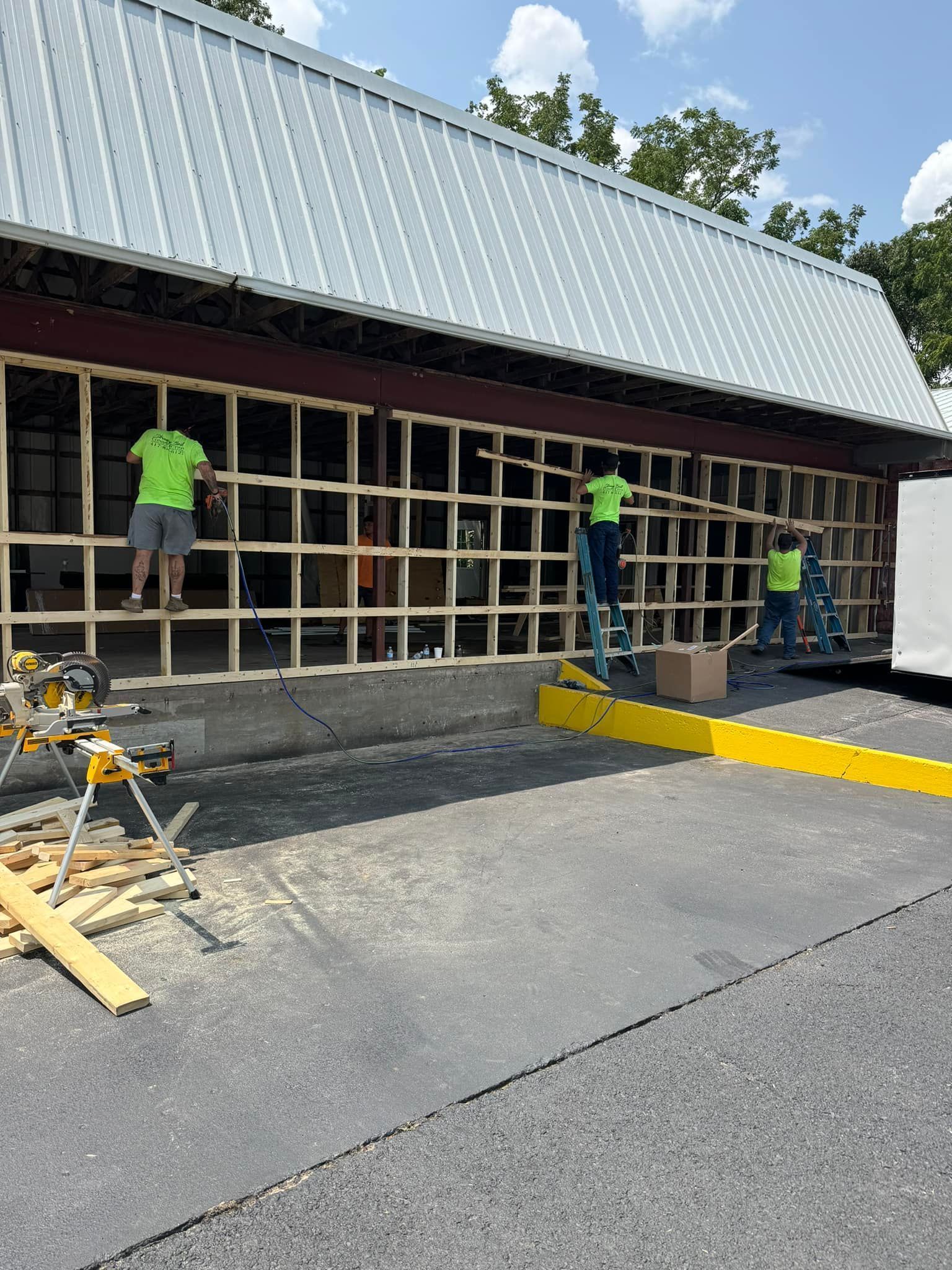 Three people in neon shirts work on a large wooden grid frame attached to the front of a building with a metal roof.