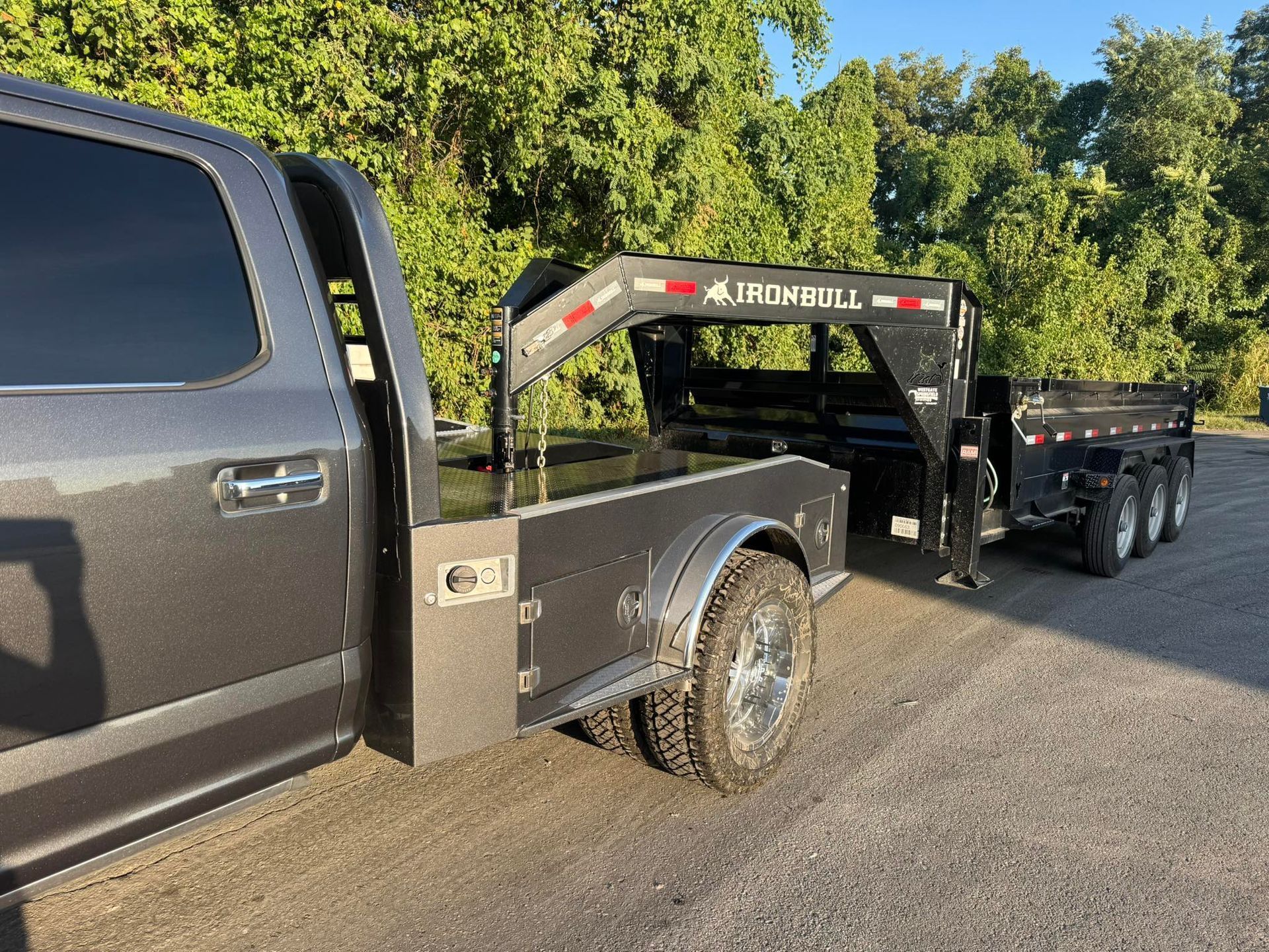 A gray pickup truck with a flatbed body is hitched to a black Iron Bull dump trailer on a dirt road near trees.