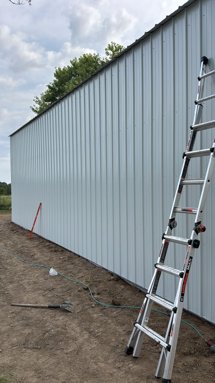 A tall, silver metal-sided building stands outdoors next to an open extension ladder leaning against its exterior wall.