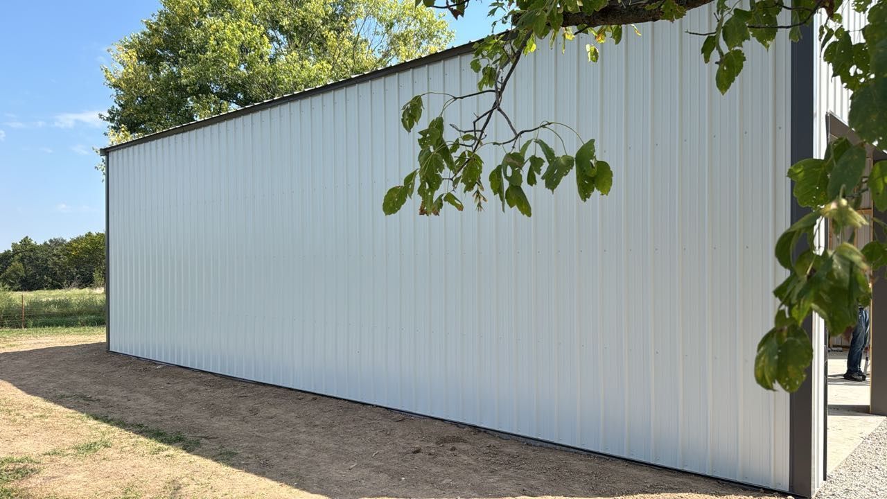 The side of a white metal pole barn sits on a dirt lot with tree branches hanging in the foreground.