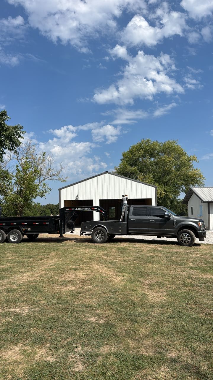 A dark grey pickup truck with a flatbed trailer attached, parked on a grassy lot in front of a white metal building.