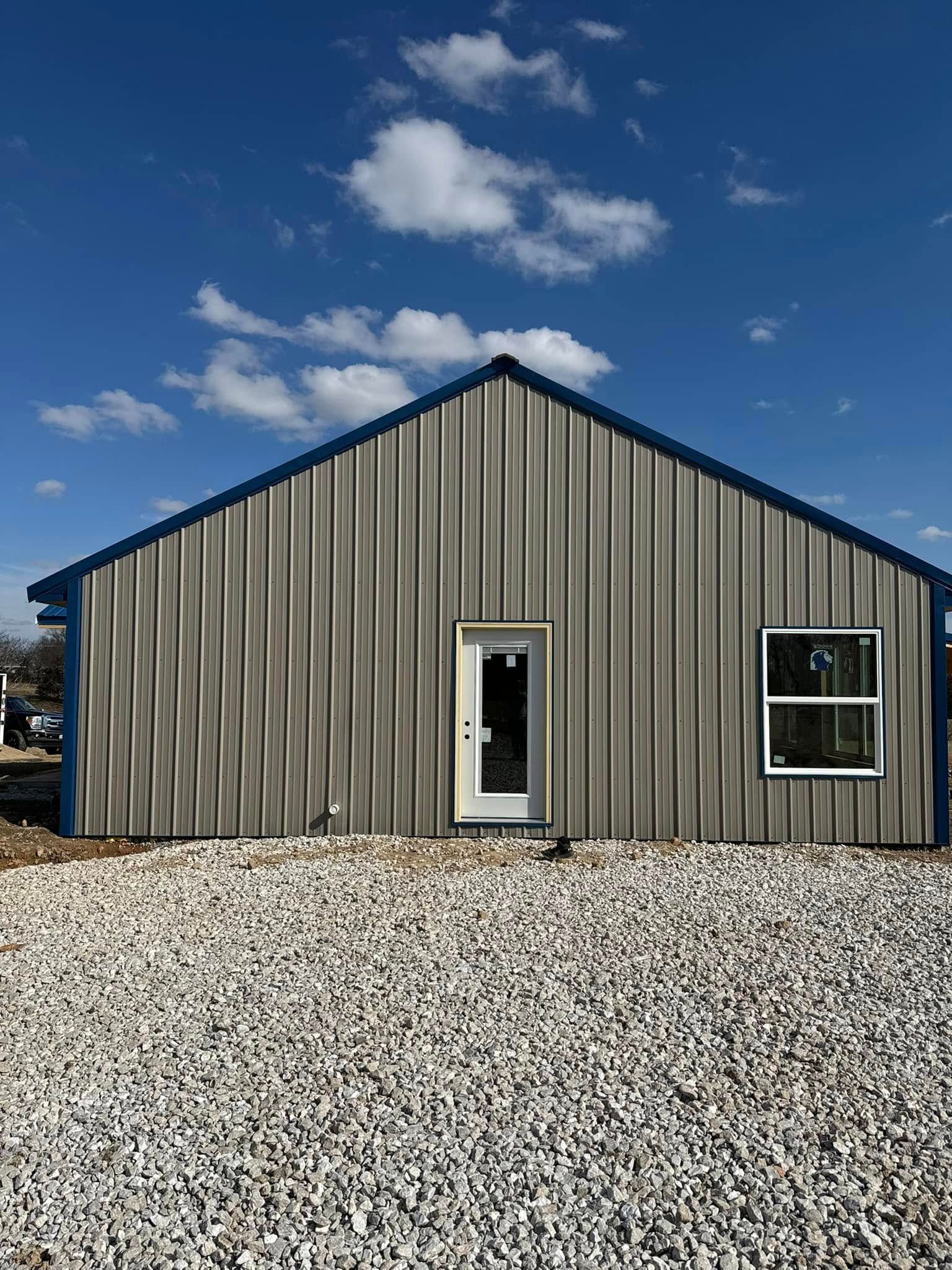 A neutral-colored metal building with a white door and a single window, set against a blue sky with gravel in the front.