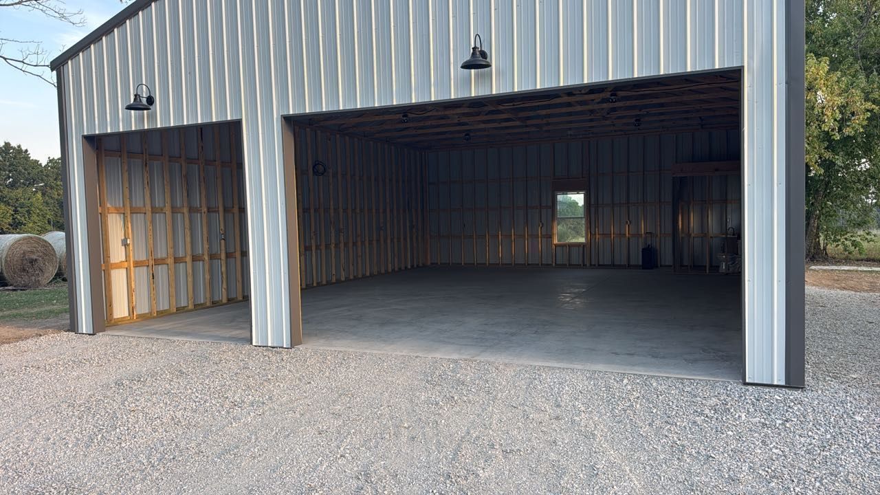 A wide, open metal-sided garage building with two large bays and a concrete floor, set on a gravel driveway.