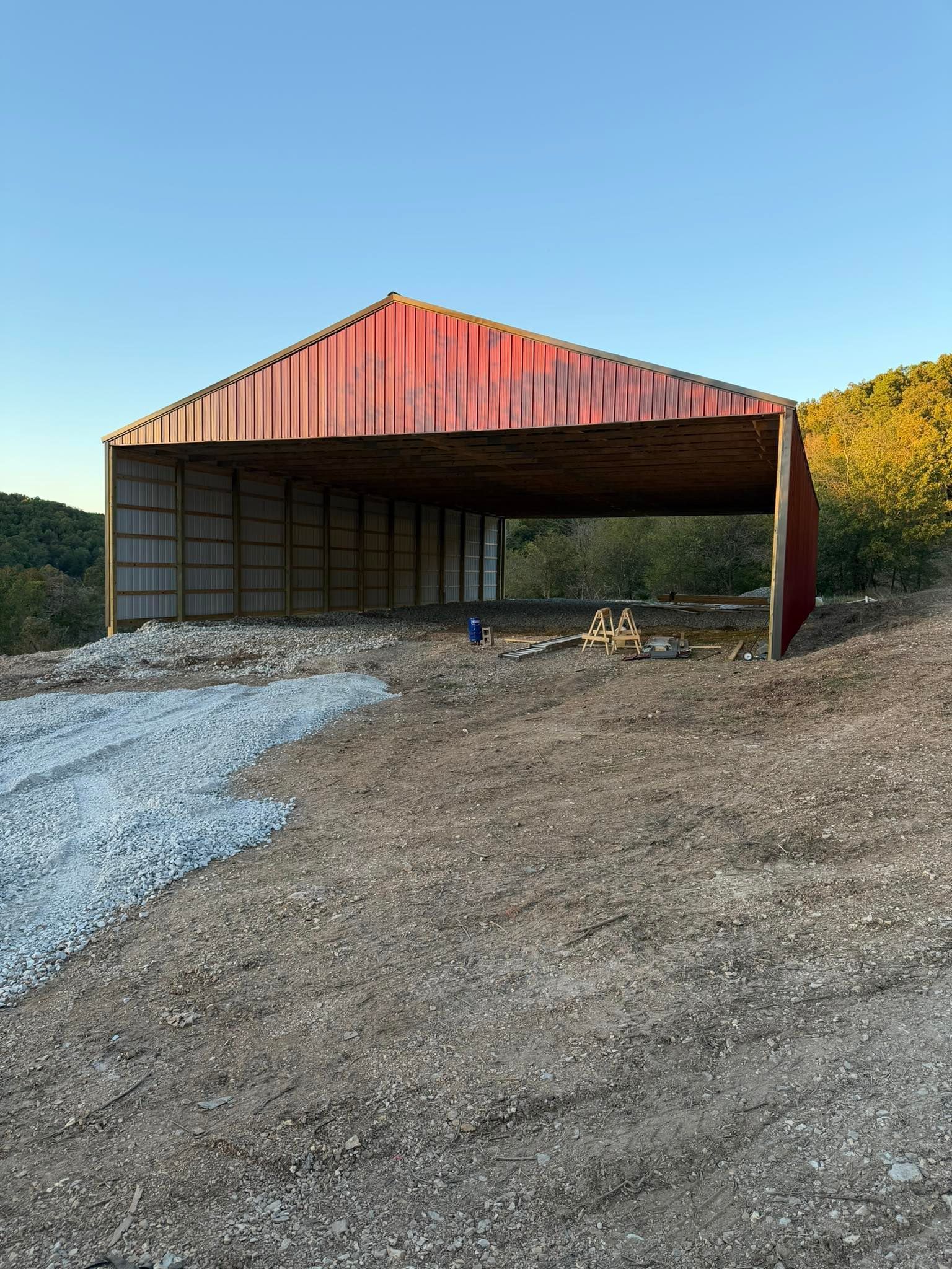 A red-roofed, open-front barn sits on a dirt and gravel lot against a backdrop of trees under a clear blue sky.