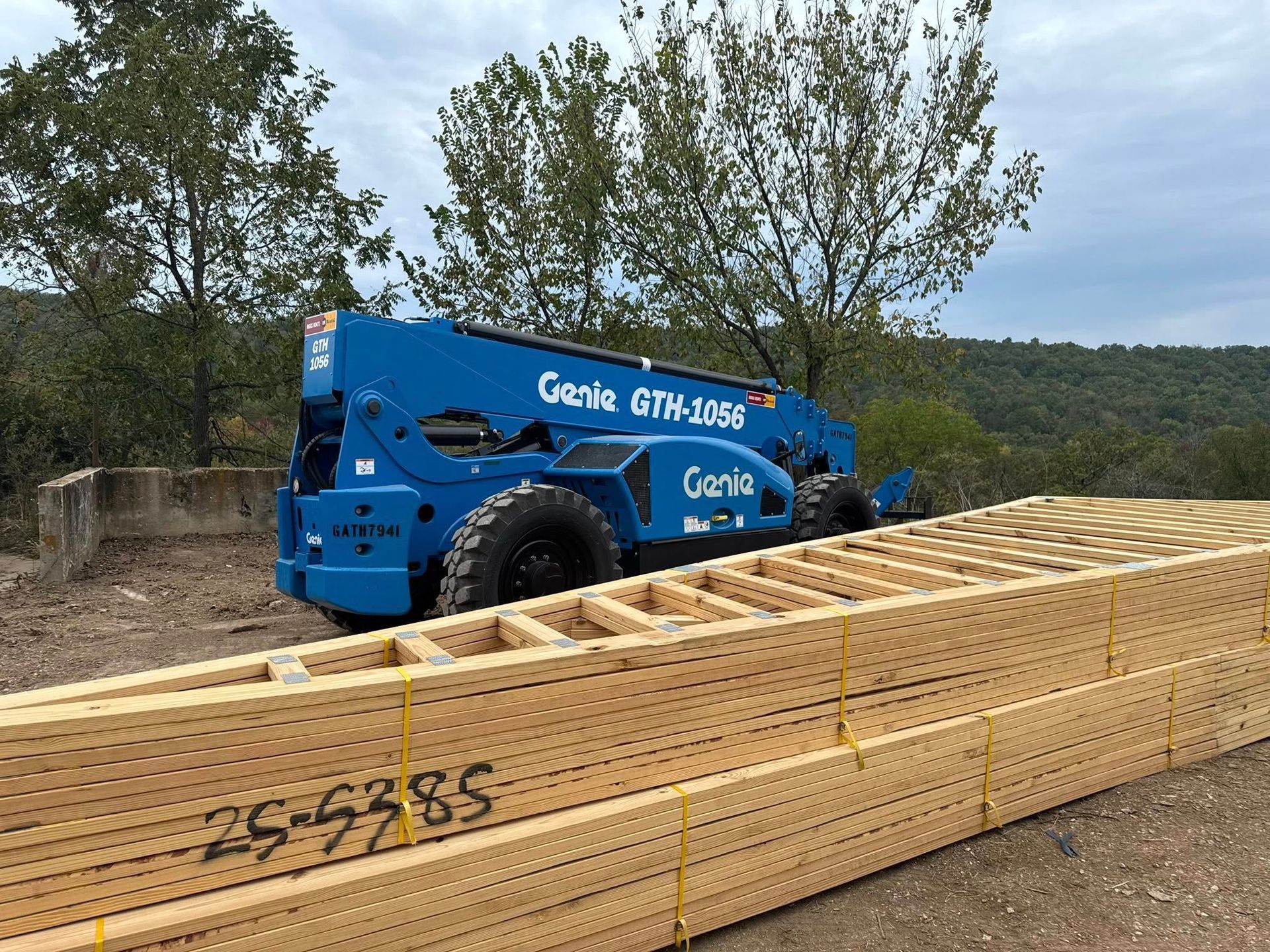 A blue Genie GTH-1056 telehandler parked behind a large stack of wooden trusses in an outdoor, rural setting.