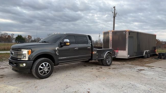 A dark gray Ford dually pickup truck towing a large, enclosed cargo trailer on a gravel lot under a cloudy sky.