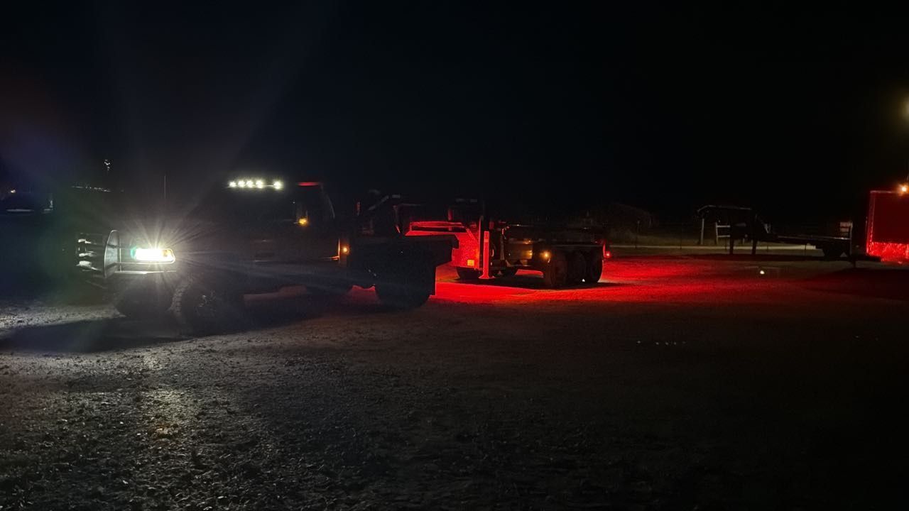 Two trucks parked on a dark, unpaved lot at night, illuminated by their headlights and red taillights.