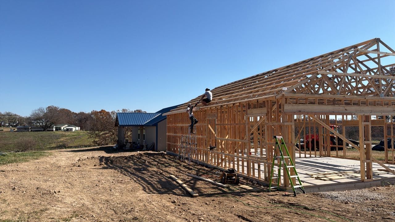 A construction worker climbs the roof trusses of a building under construction on a sunny, rural lot.