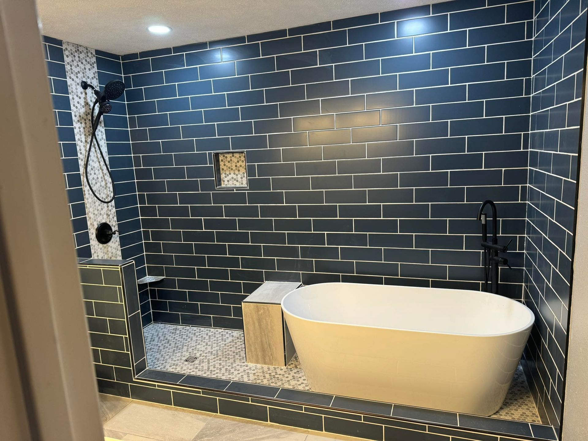 Modern bathroom with a freestanding white tub, dark blue subway tile walls, and a pebble-stone shower floor.