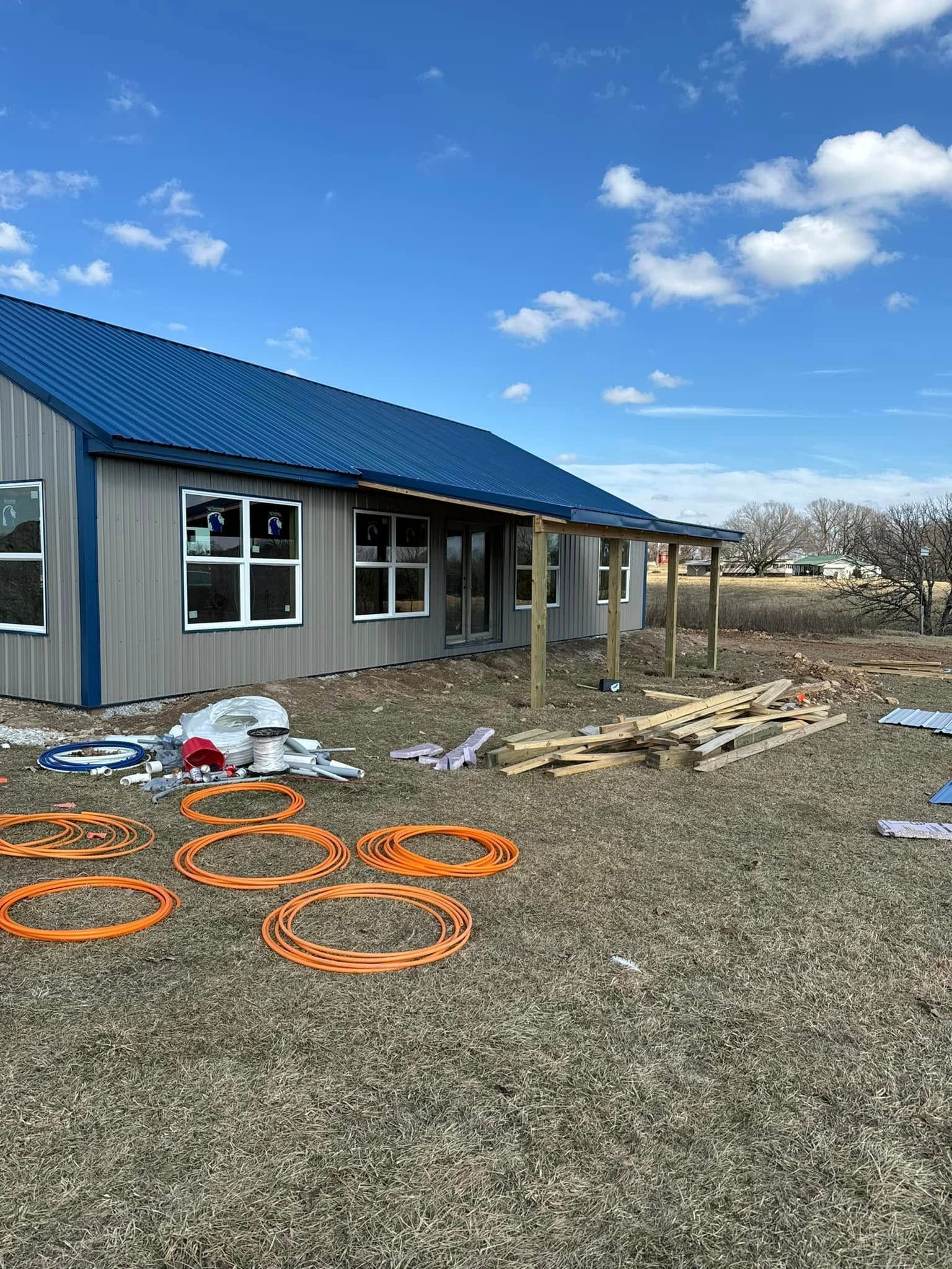 A gray metal building with a blue roof sits on a grassy lot, featuring an unfinished wooden porch and orange hoses.