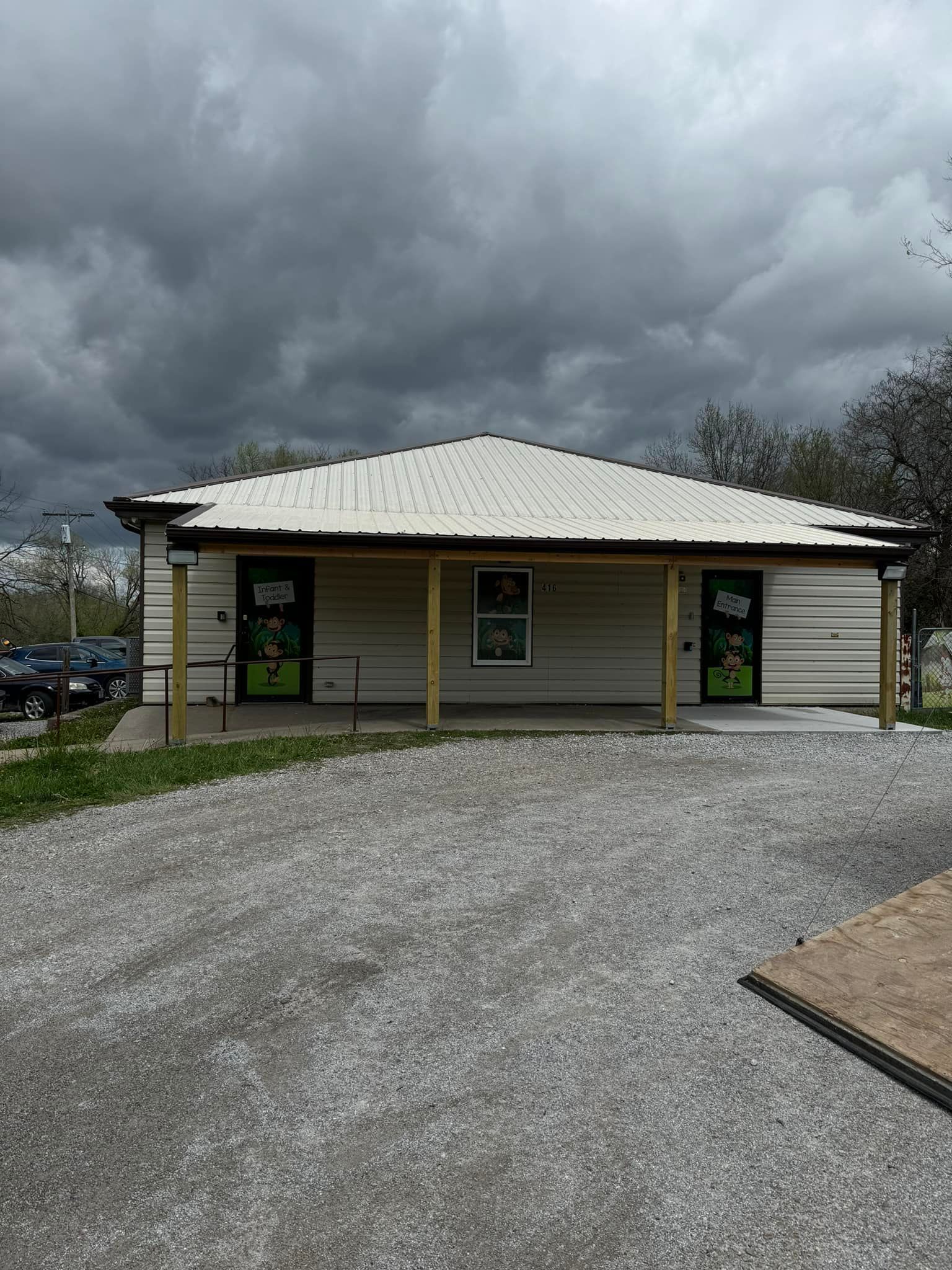 A low-set building with a covered porch and white siding under a cloudy sky, with a gravel parking area in the foreground.