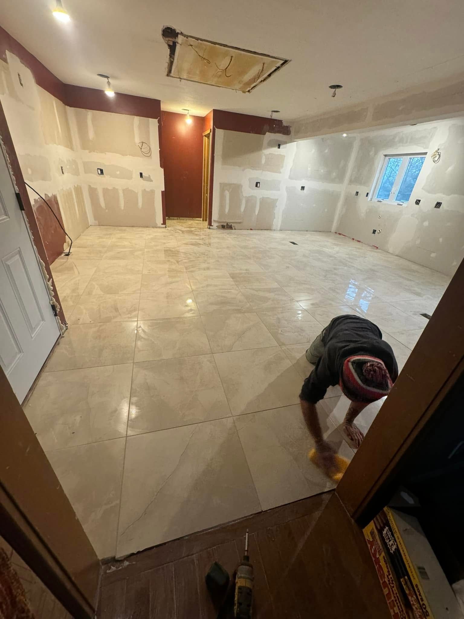 A person installing large, light-colored square tiles on the floor of a room with unfinished drywall walls.