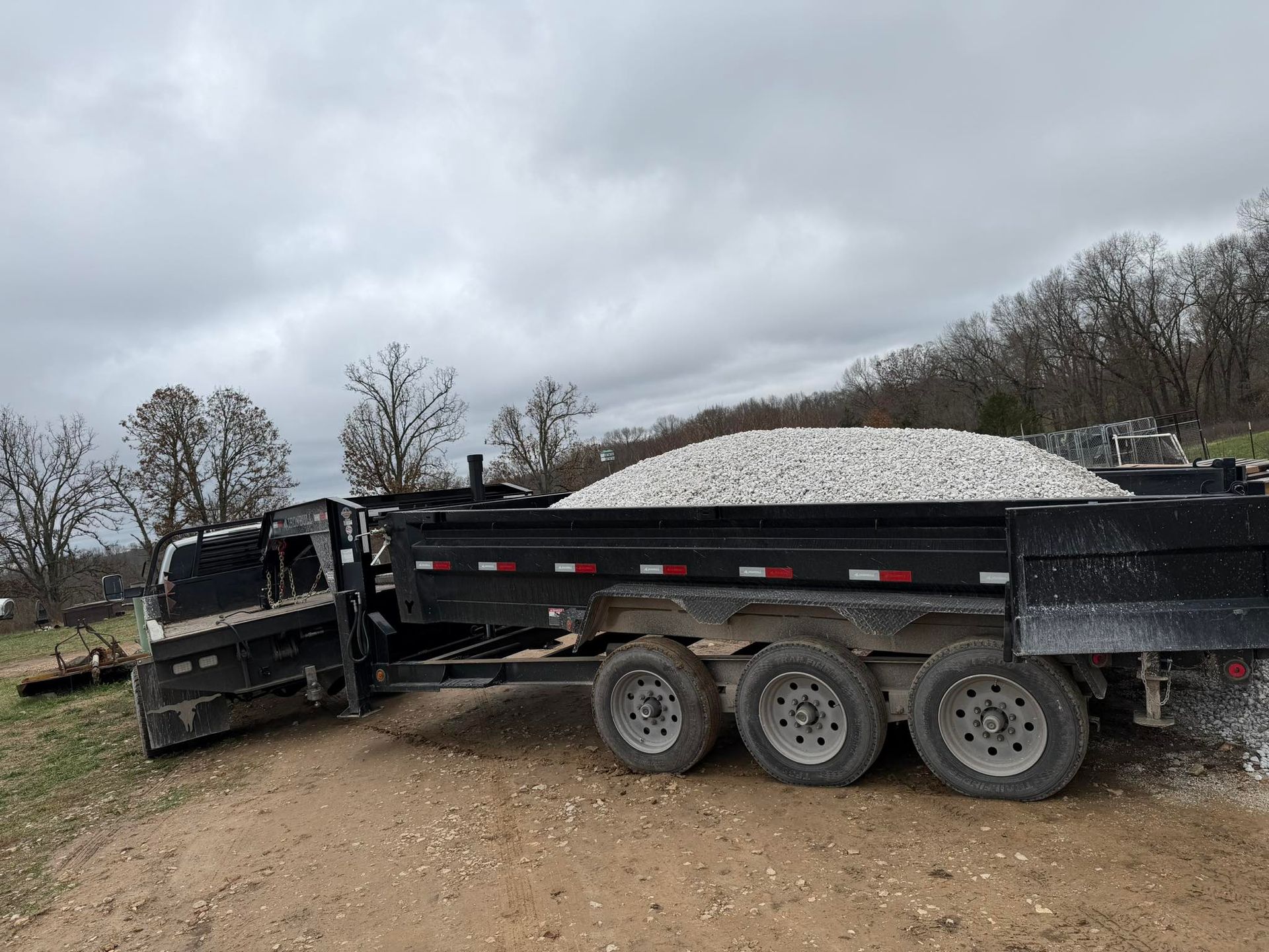 A black dump trailer filled with white gravel sits on a dirt driveway under a cloudy sky.