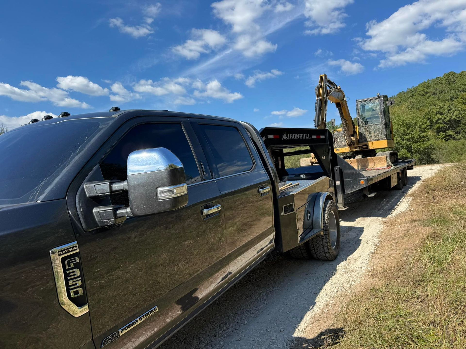 A black flatbed truck hauls a piece of construction equipment on a trailer along a dirt road under a sunny blue sky.