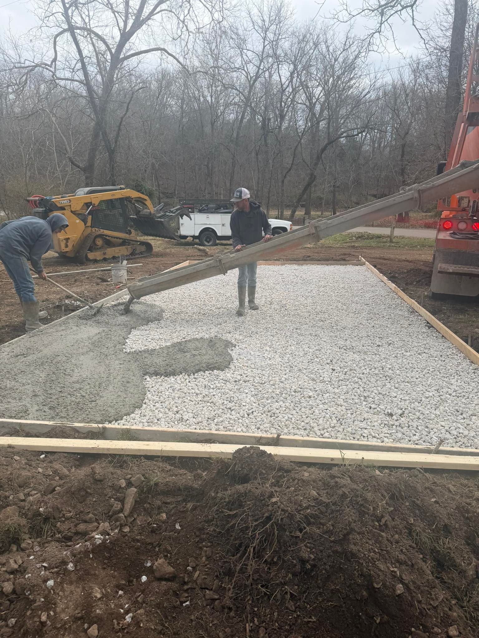 Workers pour concrete from a chute onto a gravel-filled wooden form in a wooded area.