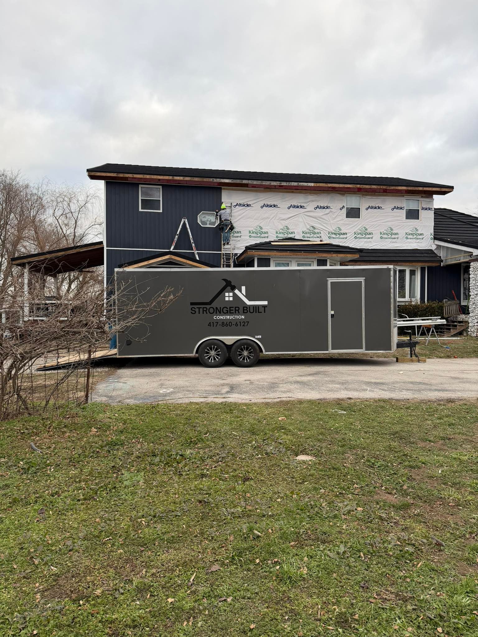 A charcoal grey cargo trailer with a white roofing company logo parked in front of a house under construction.