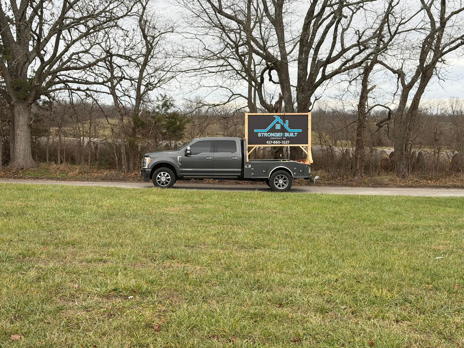 A dark grey pickup truck parked in a grassy field with a large business sign mounted in its truck bed.