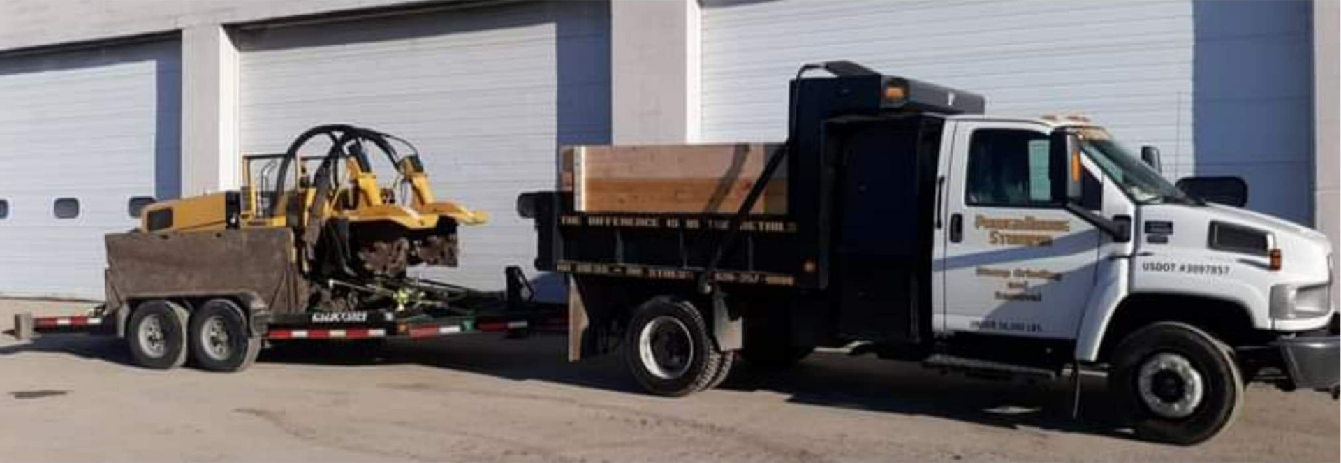 A white dump truck hauling a yellow construction machine on a flatbed trailer in front of a building with white doors.
