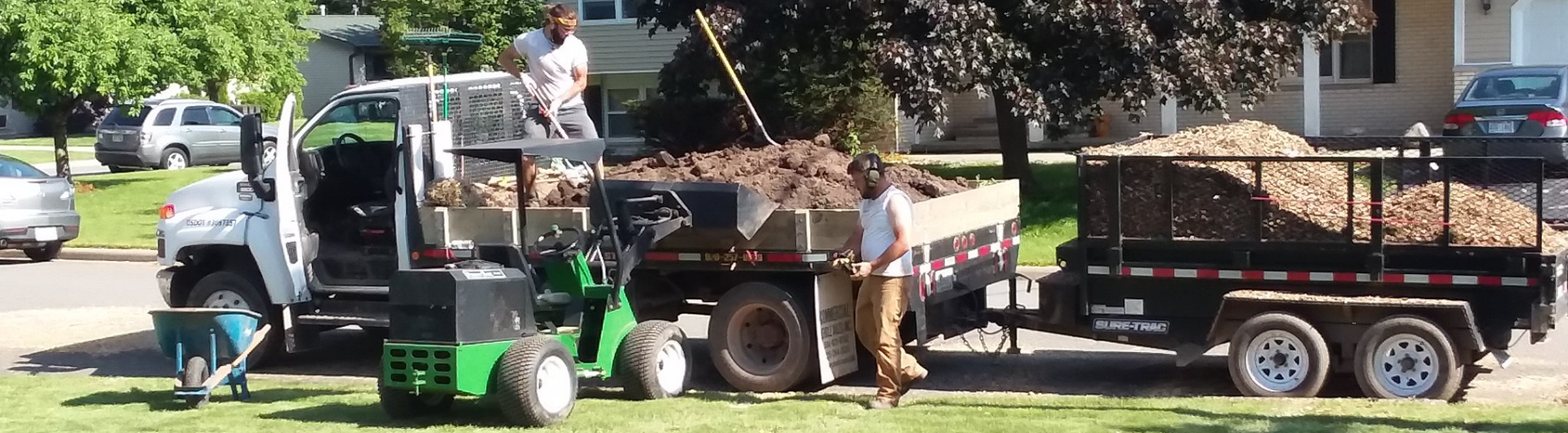 Two people work on a landscaping project, loading wood chips into a dump truck and trailer parked on a suburban street.