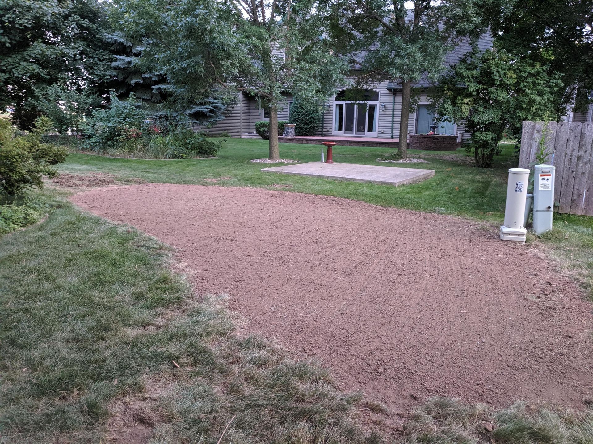 A residential backyard featuring a patch of freshly leveled reddish soil with a house and patio area in the background.