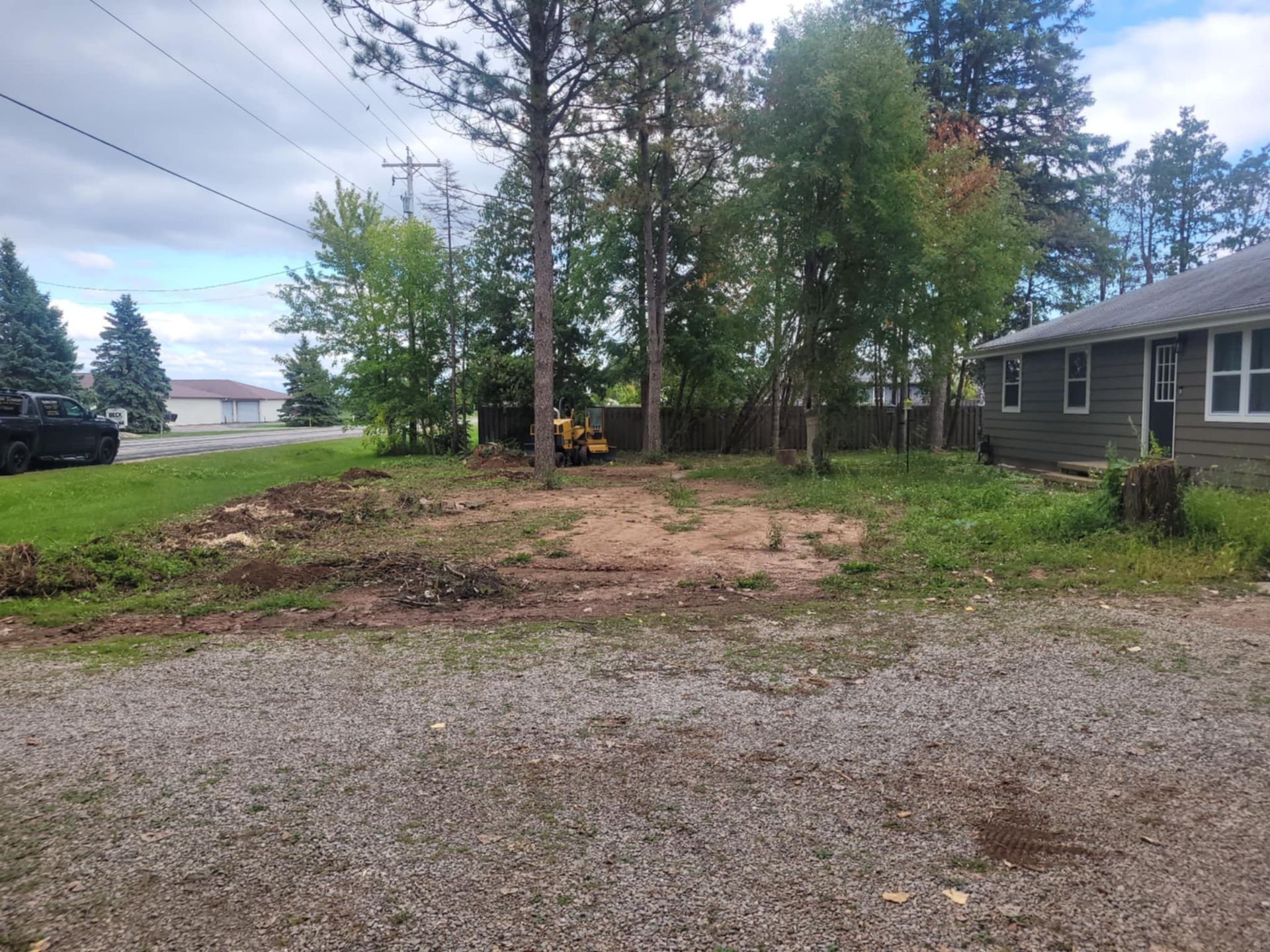 A dirt clearing with a small piece of construction equipment behind a grey house and trees on a gravel lot.