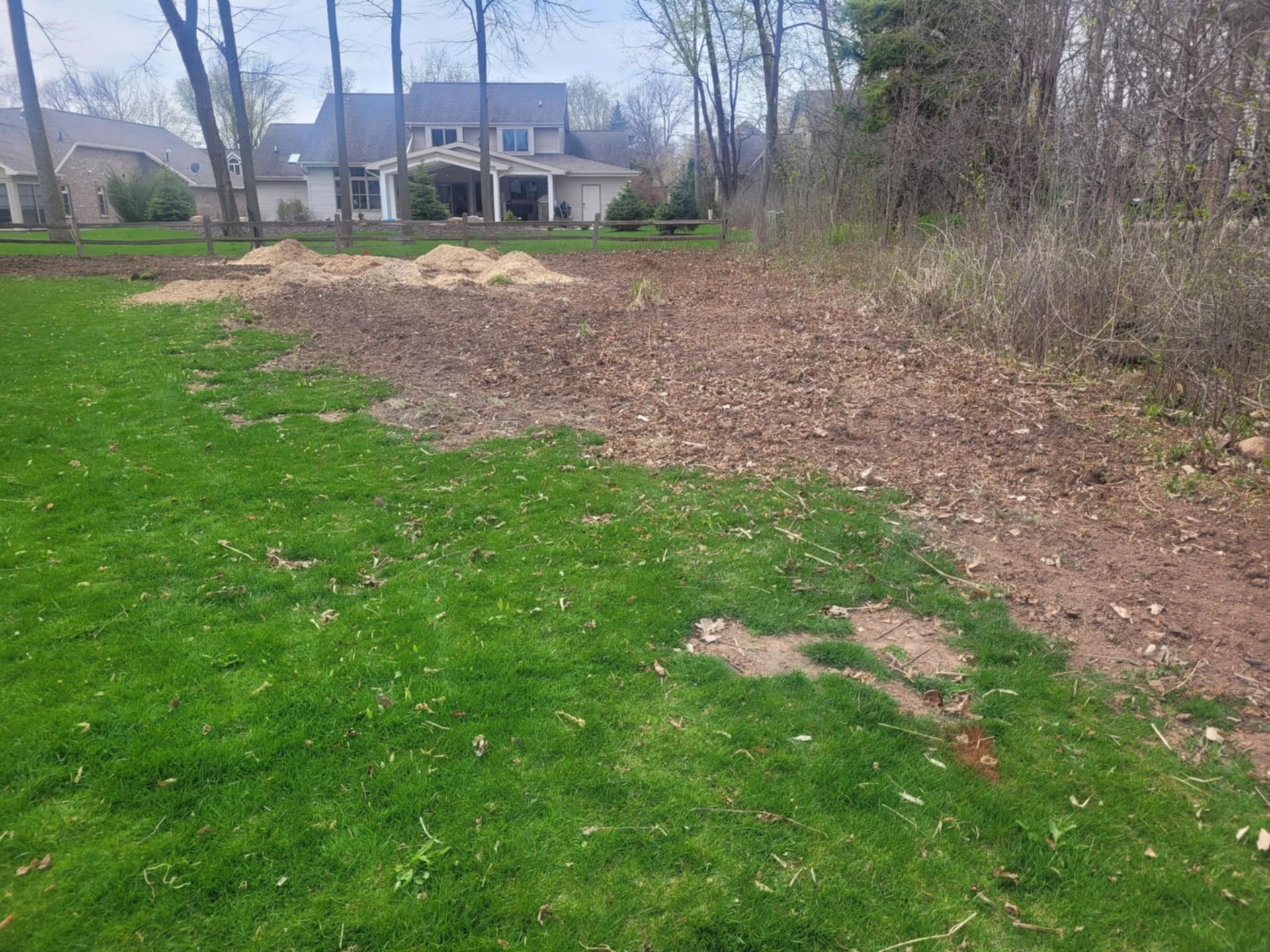 A view of a backyard featuring a lush green lawn transitioning into an area of cleared soil, wood chips, and mulch.