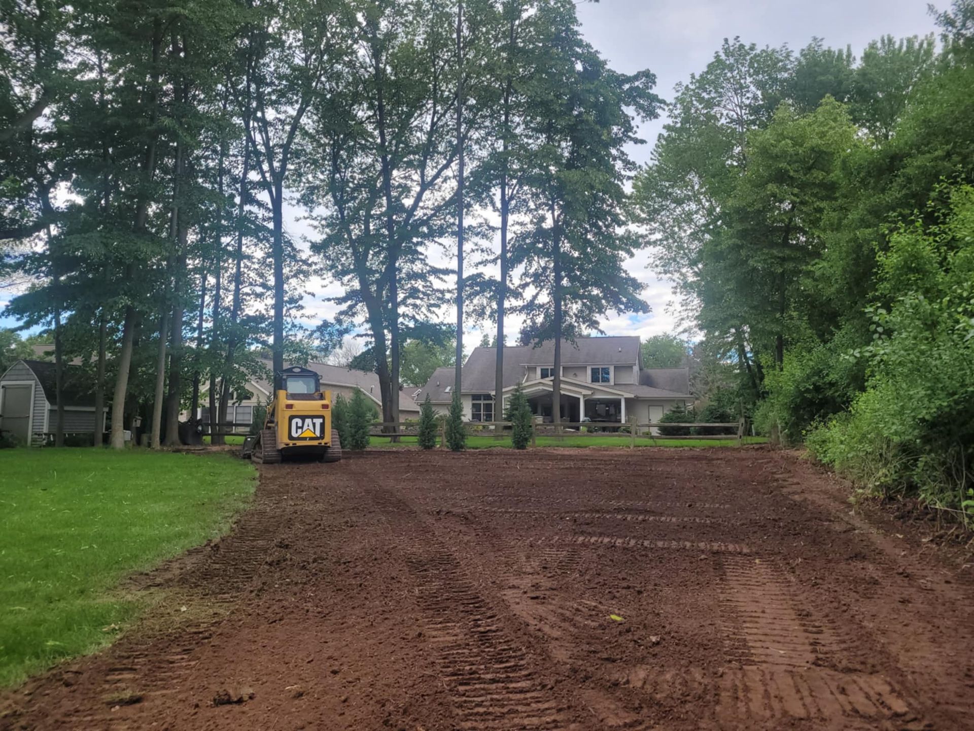 A yellow Caterpillar skid steer clears a large, bare patch of dirt in a grassy residential yard bordered by tall trees.