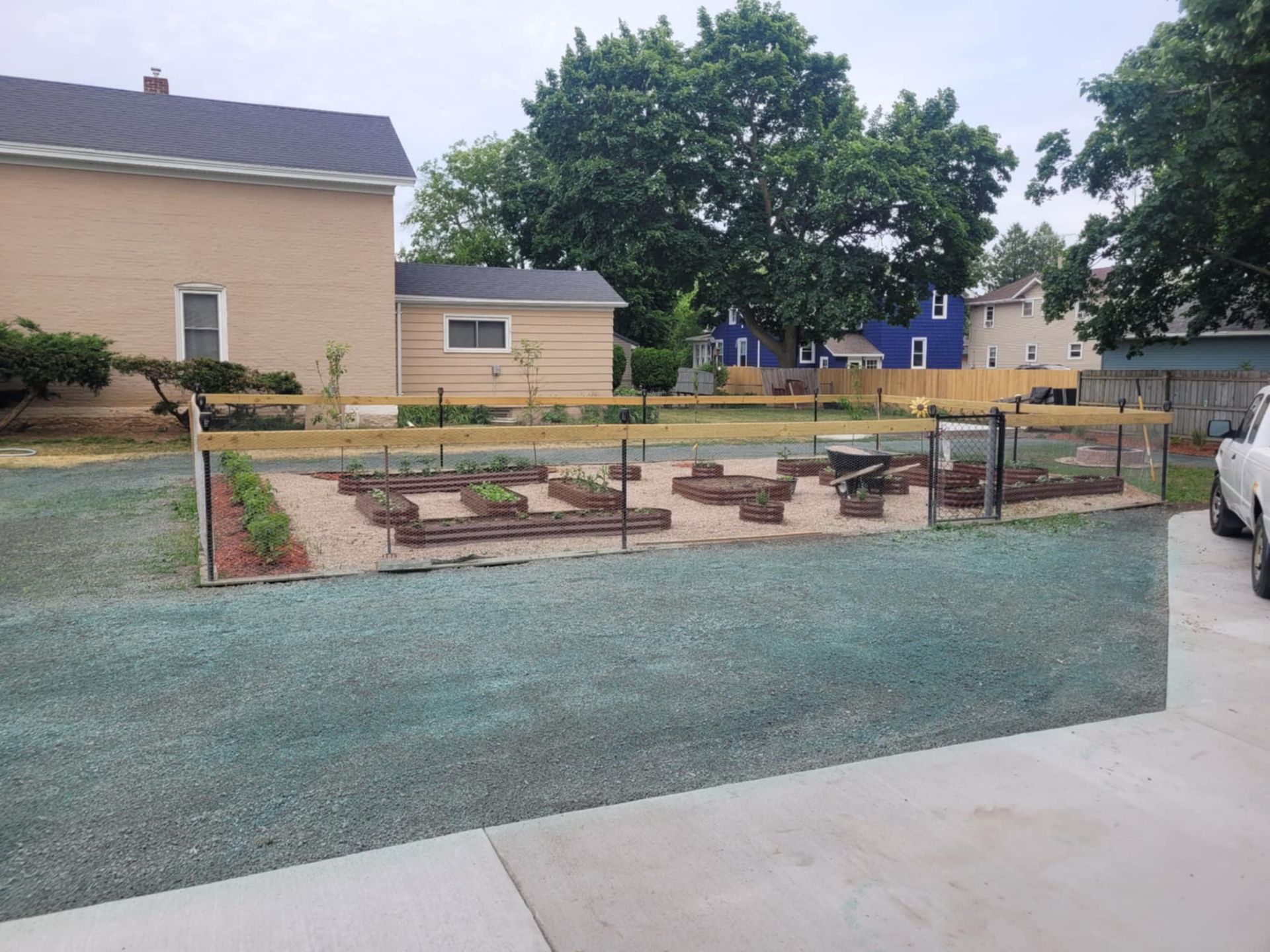 A fenced garden with raised beds, gravel paths, and small plants, situated in a residential yard near a tan house.