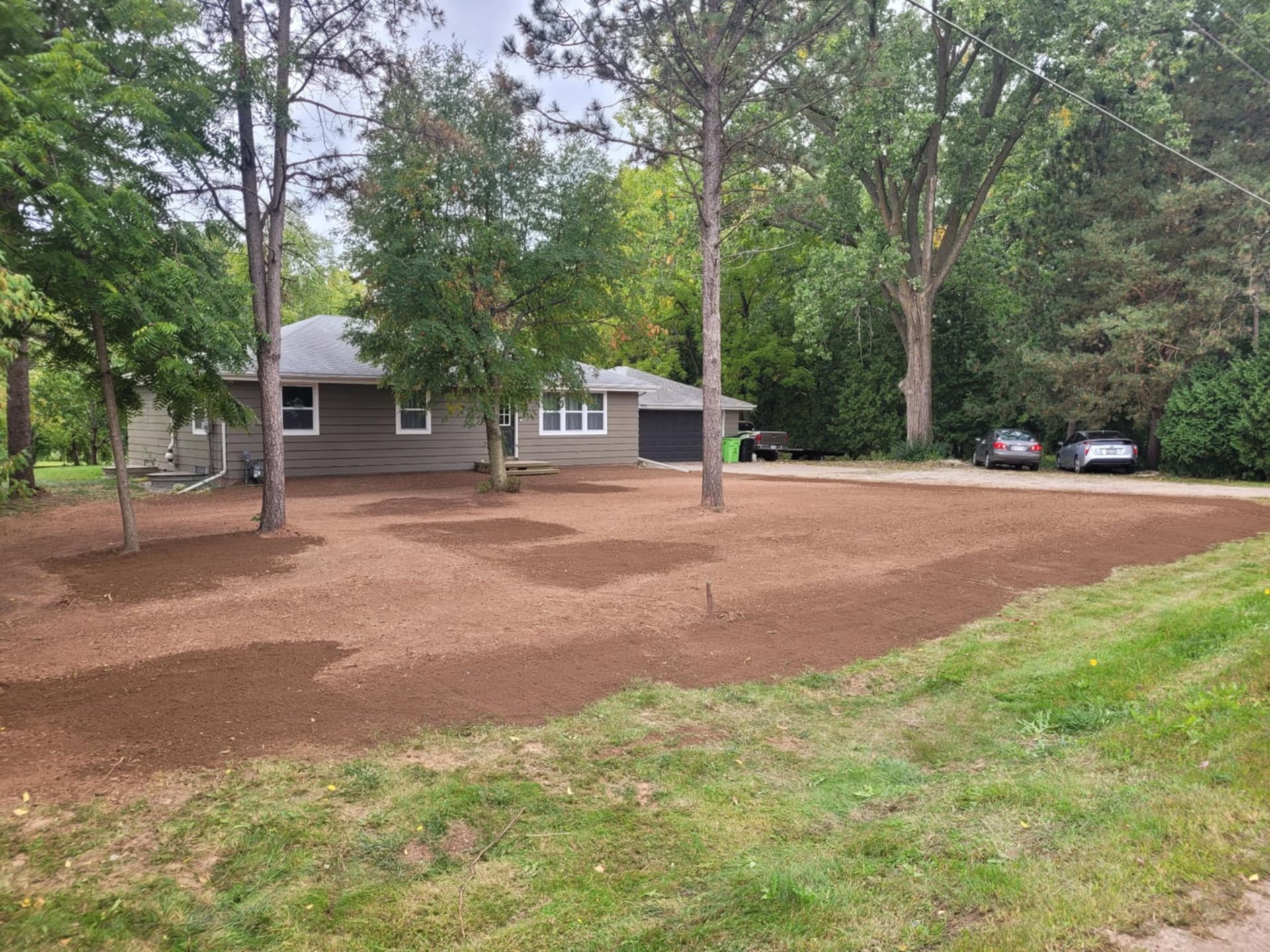 A grey house behind a large, freshly prepared dirt lawn surrounded by mature trees.
