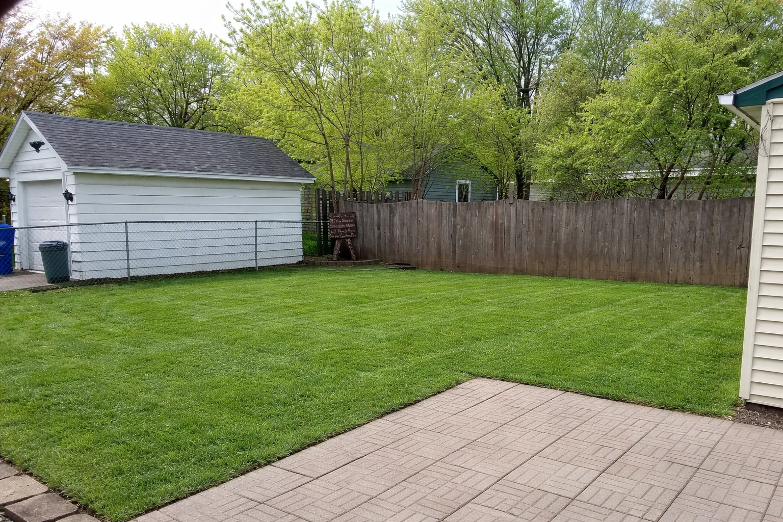 A backyard with a stone patio in the foreground, a manicured green lawn, a white detached garage, and a wooden fence.