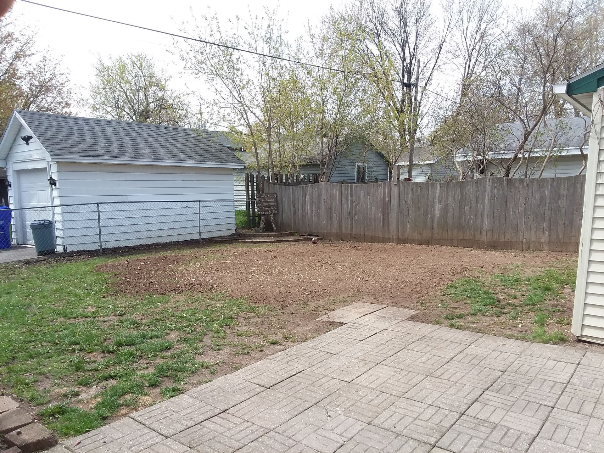 A view of a backyard with a stone patio in the foreground, a grassy area, a white garage, and a wooden fence.