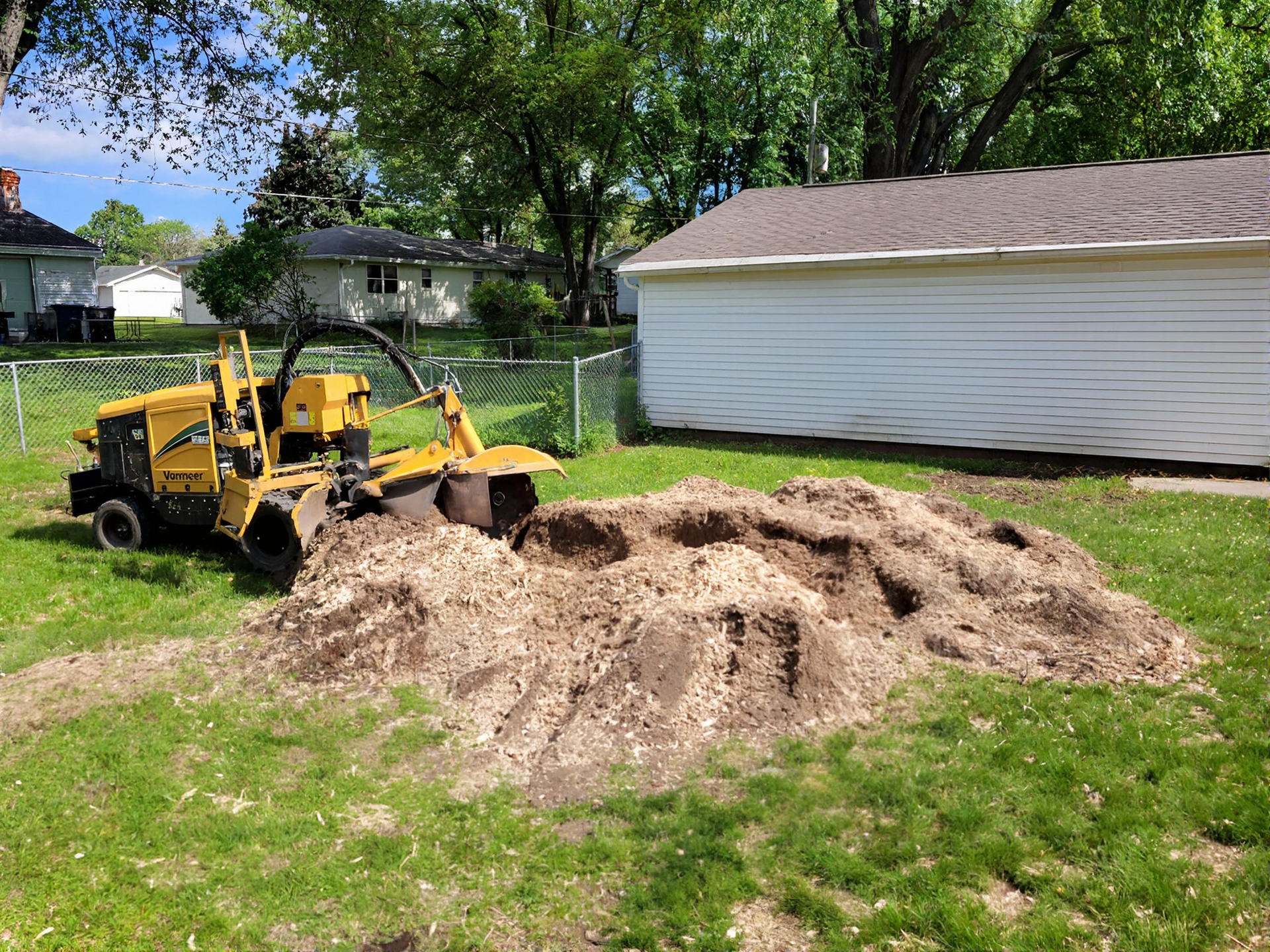 A yellow stump grinder cuts into a tree stump, throwing wood chips in an outdoor residential setting.