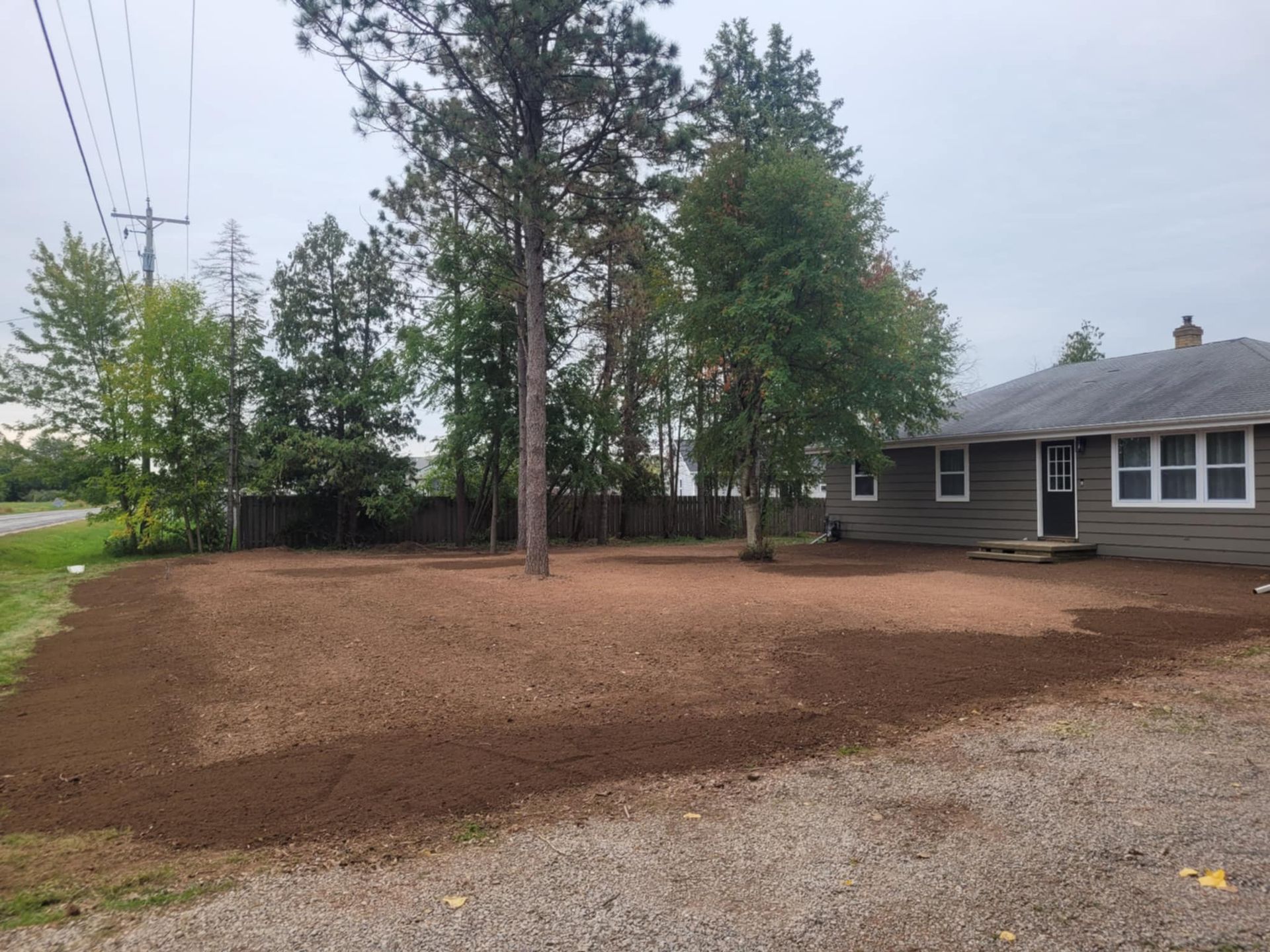 A residential yard with a house on the right, featuring a large area of freshly tilled, dark brown soil and tall trees.