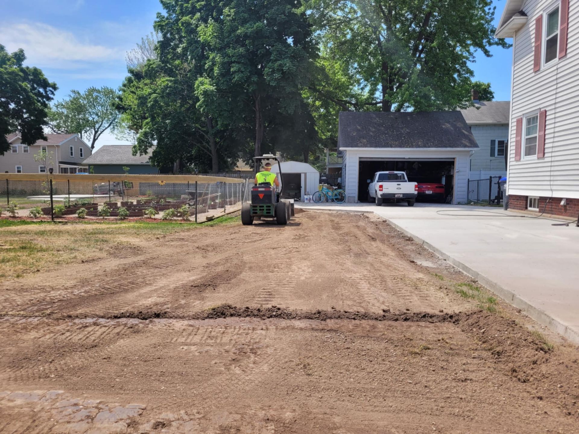 A construction worker on a compact tractor levels dirt in a yard next to a white house and garage on a sunny day.