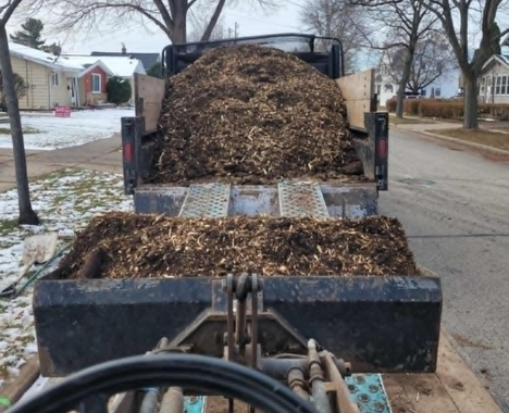 A close-up view from a piece of construction equipment, showing a metal bucket filled with wood chips in front of a truck.