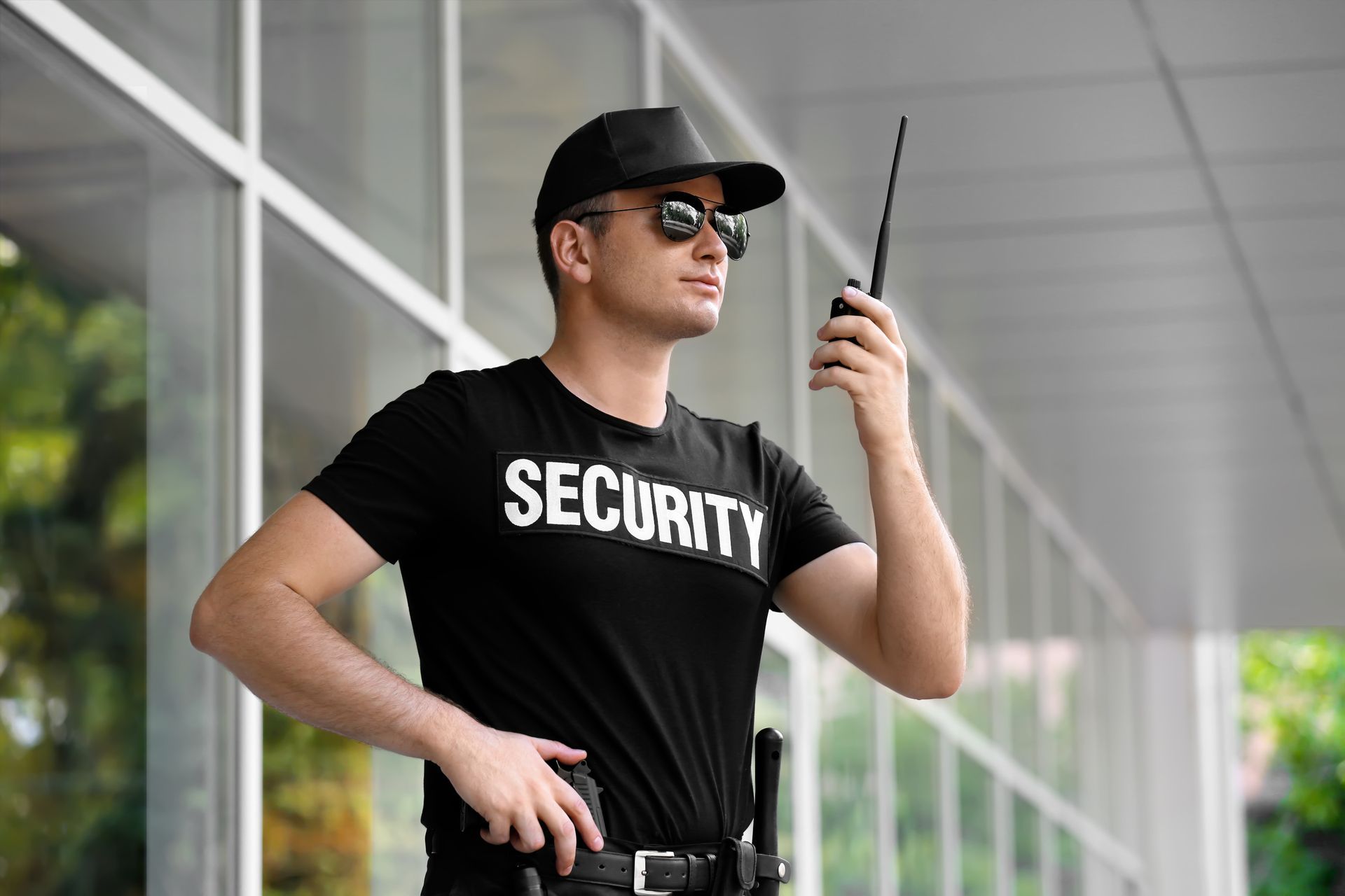 A security guard in a black uniform and sunglasses holds a walkie-talkie outside a building with glass walls.