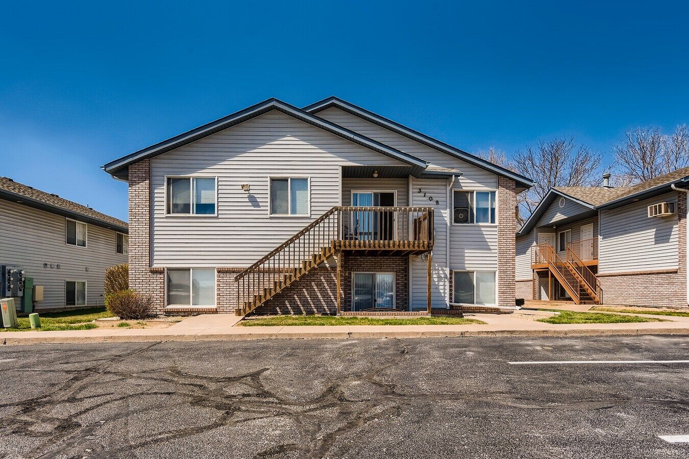 Two-story light gray apartment building with wooden stairs and a small balcony against a blue sky.
