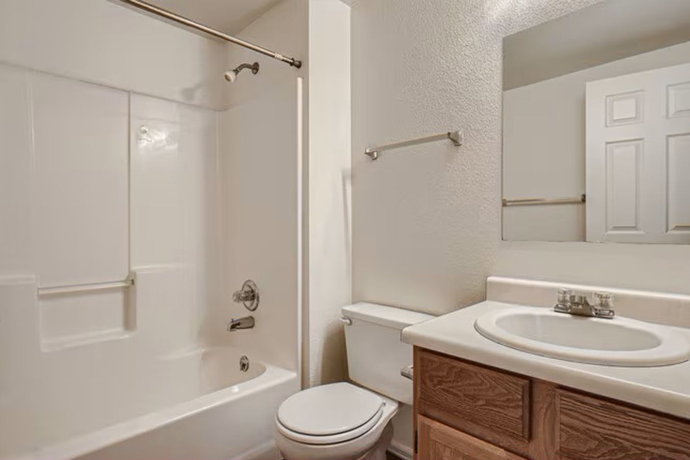 Bathroom with white tub/shower, toilet, vanity with sink, and mirror. Beige walls, light brown cabinets.