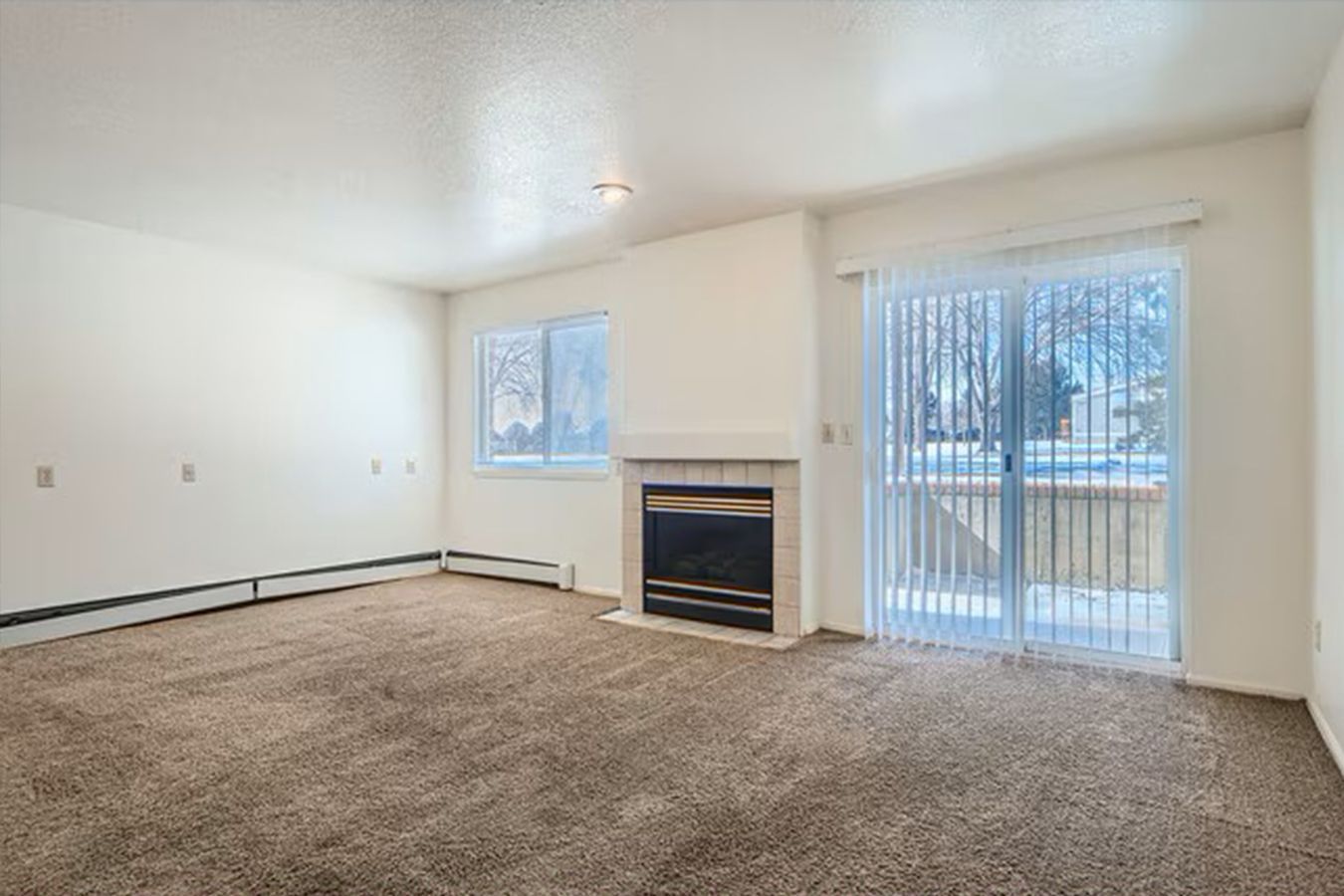 Empty living room with fireplace, sliding glass door, and window; beige carpet and walls.