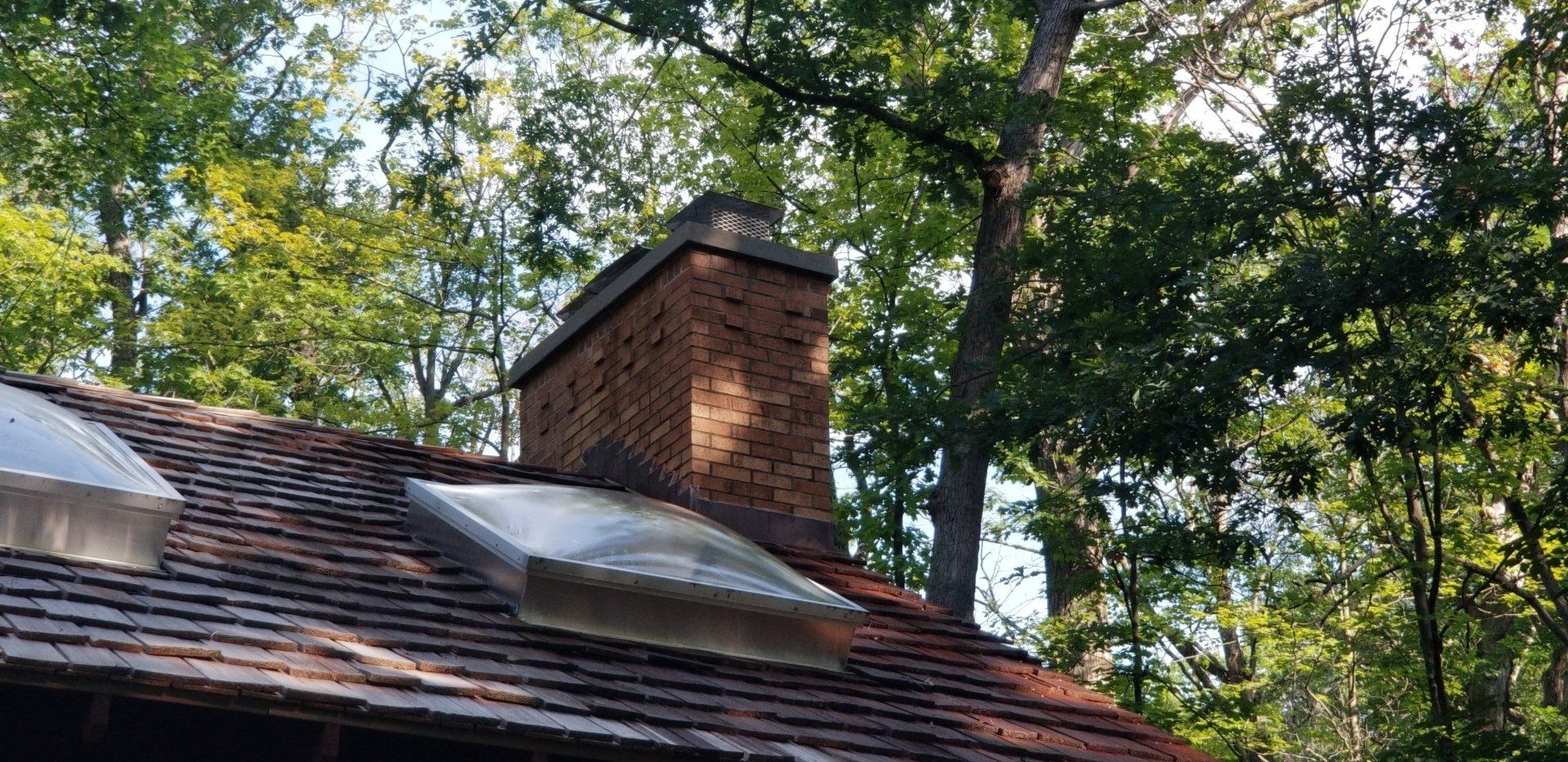 A brick chimney is on the roof of a house surrounded by trees.