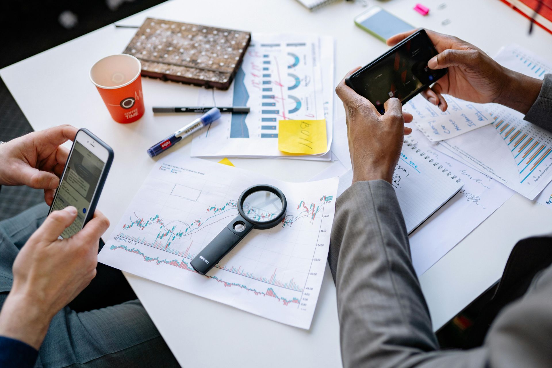 People looking at documents and phones at a table with charts, a magnifying glass, and a coffee cup.