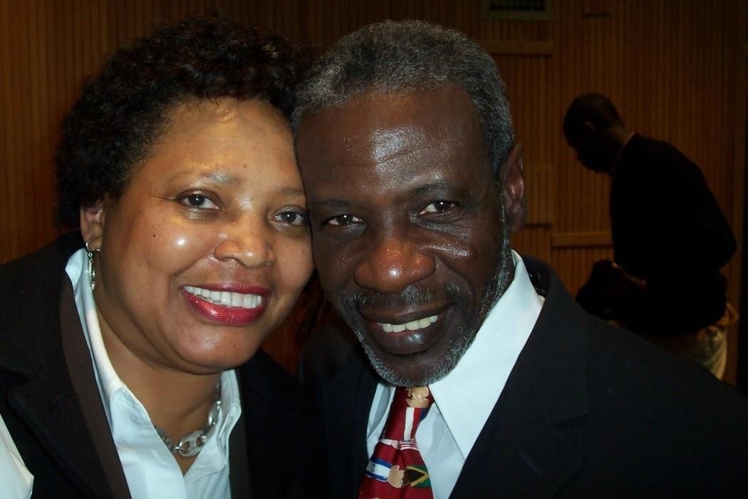 Two people smiling close to the camera. Man in suit, woman in white blouse and jacket. Neutral background, indoors.