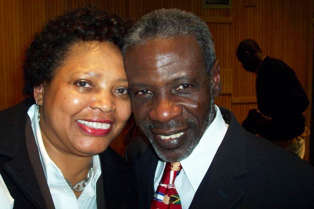 Husband and wife smiling at the camera, close up. The man wears a suit and tie. The woman has short hair.